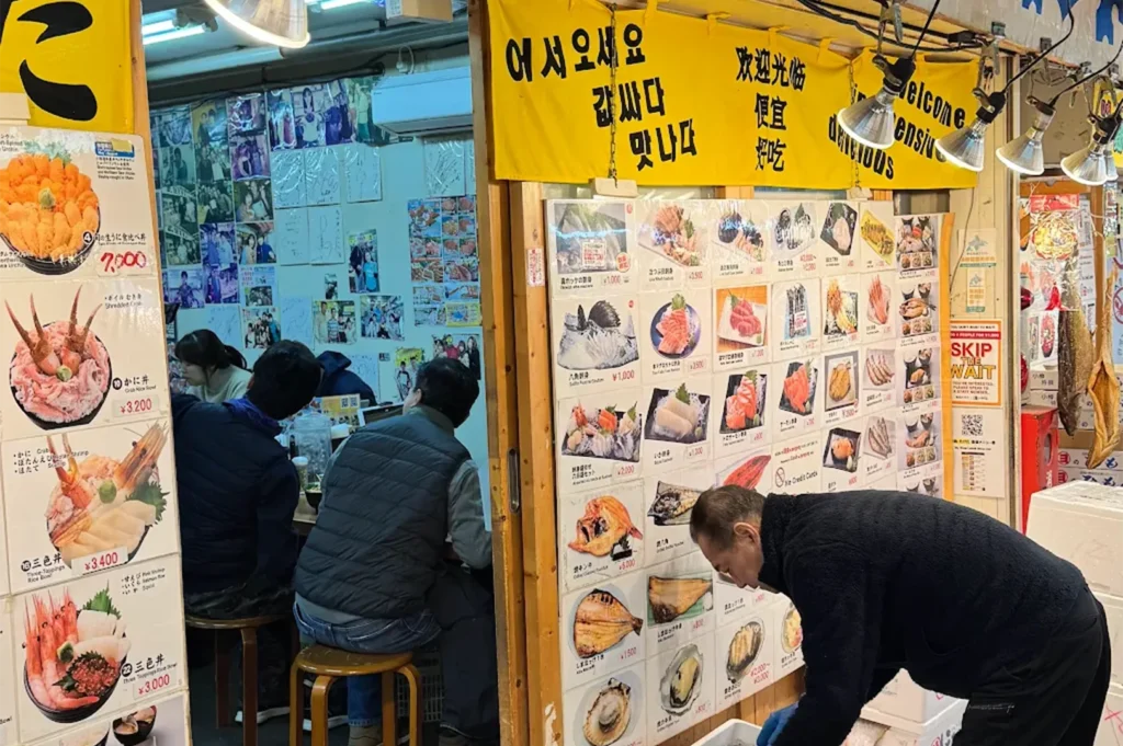 Interior of Ajidokoro Takeda in Otaru's Sankaku Market, showing customers seated at the bar and a wall covered in photos and seafood bowl menus.