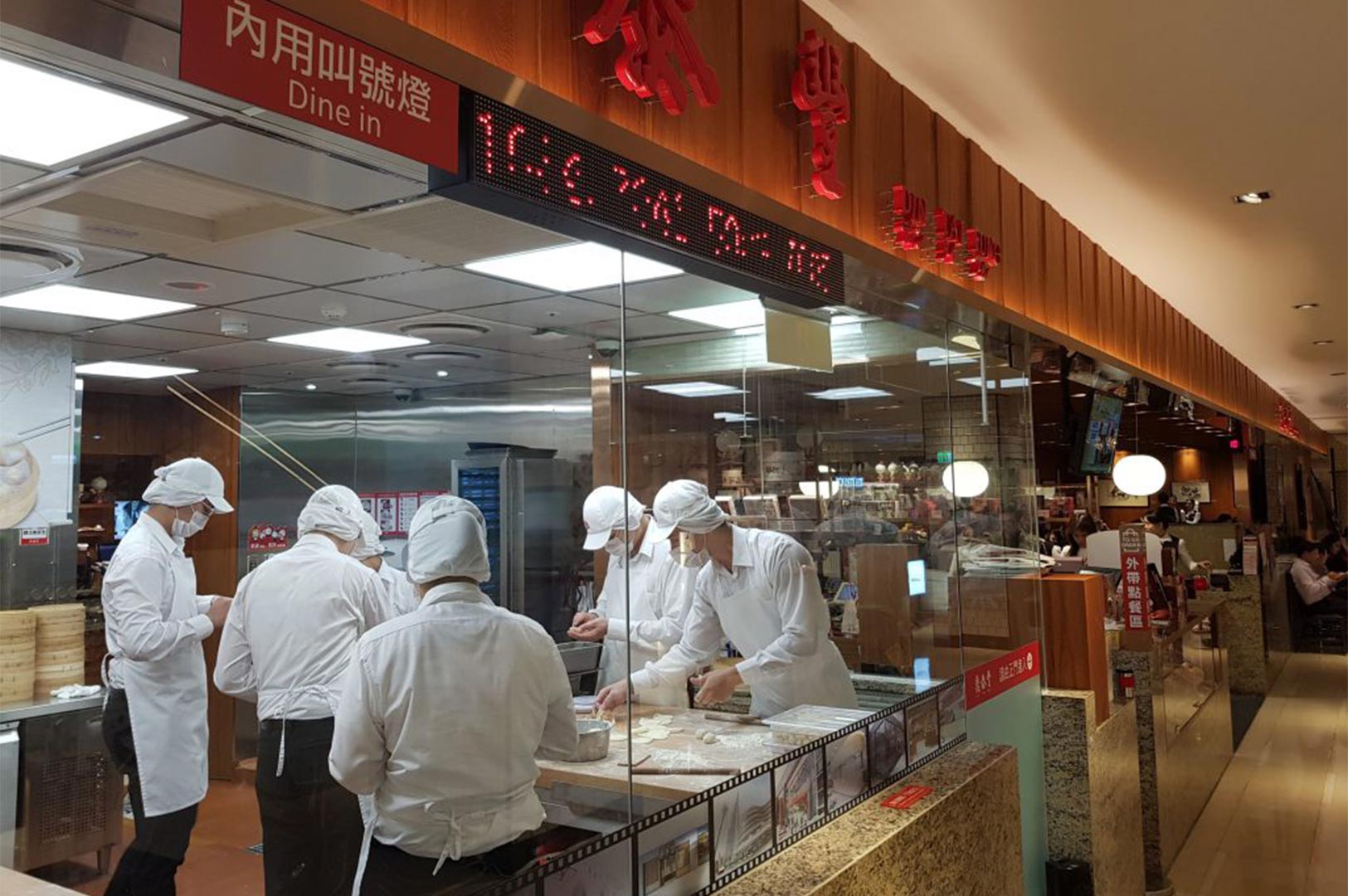 Professional chefs in white uniforms and masks meticulously hand-folding xiaolongbao dumplings behind a glass window at a Din Tai Fung restaurant.