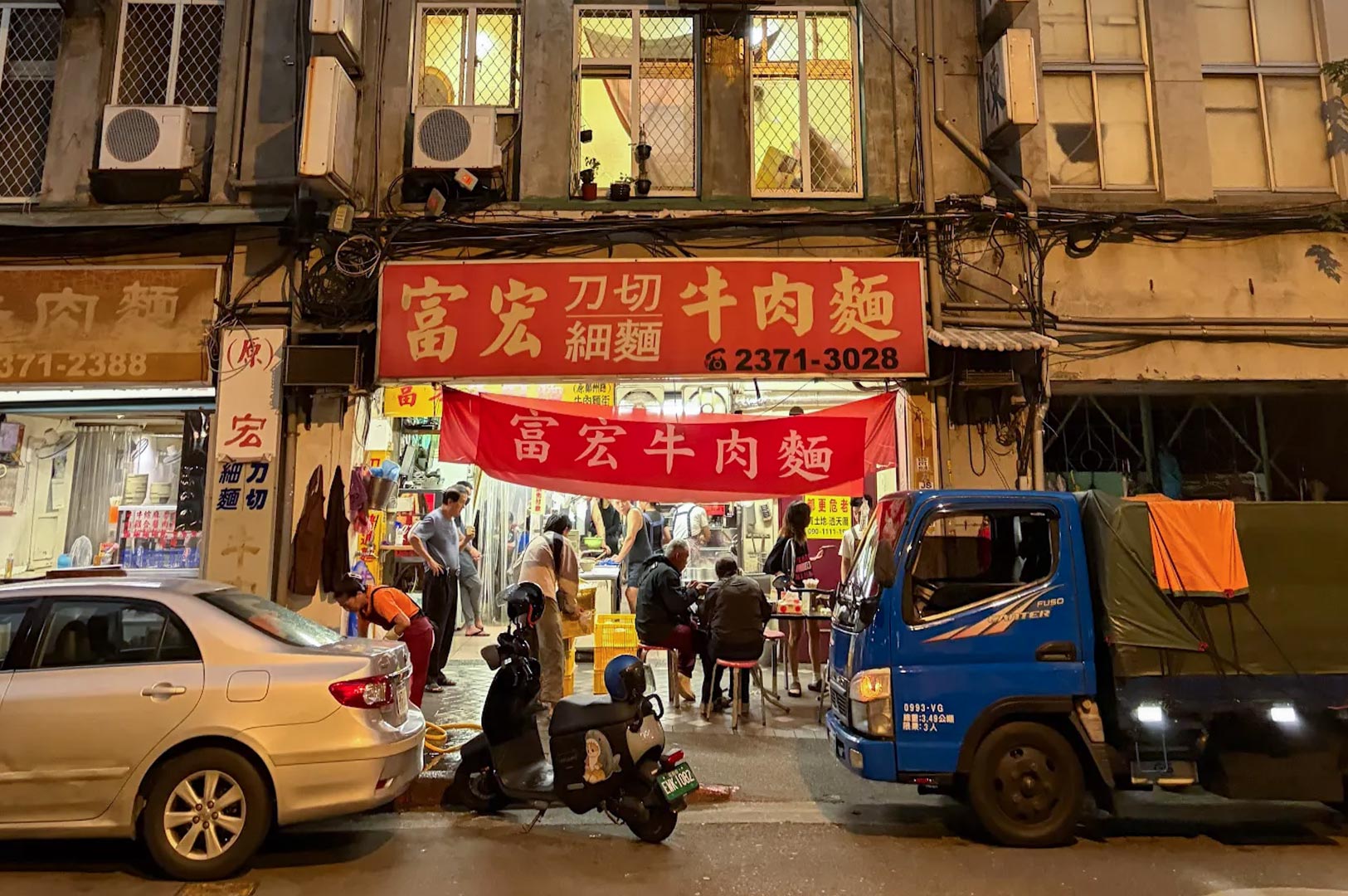 A vibrant night view of Fuhong Beef Noodles in Taipei, featuring a bright red sign, customers dining at outdoor tables, and a blue delivery truck parked in front.