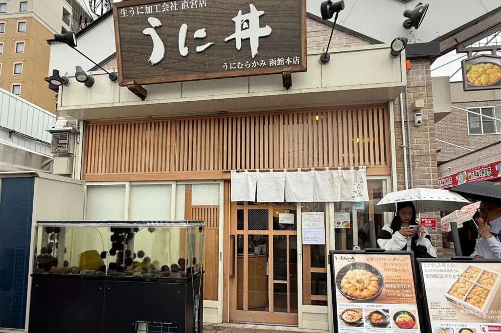 The exterior of Uni Murakami main store in Hakodate, featuring a wooden "Uni-don" sign and a glass tank filled with live sea urchins.