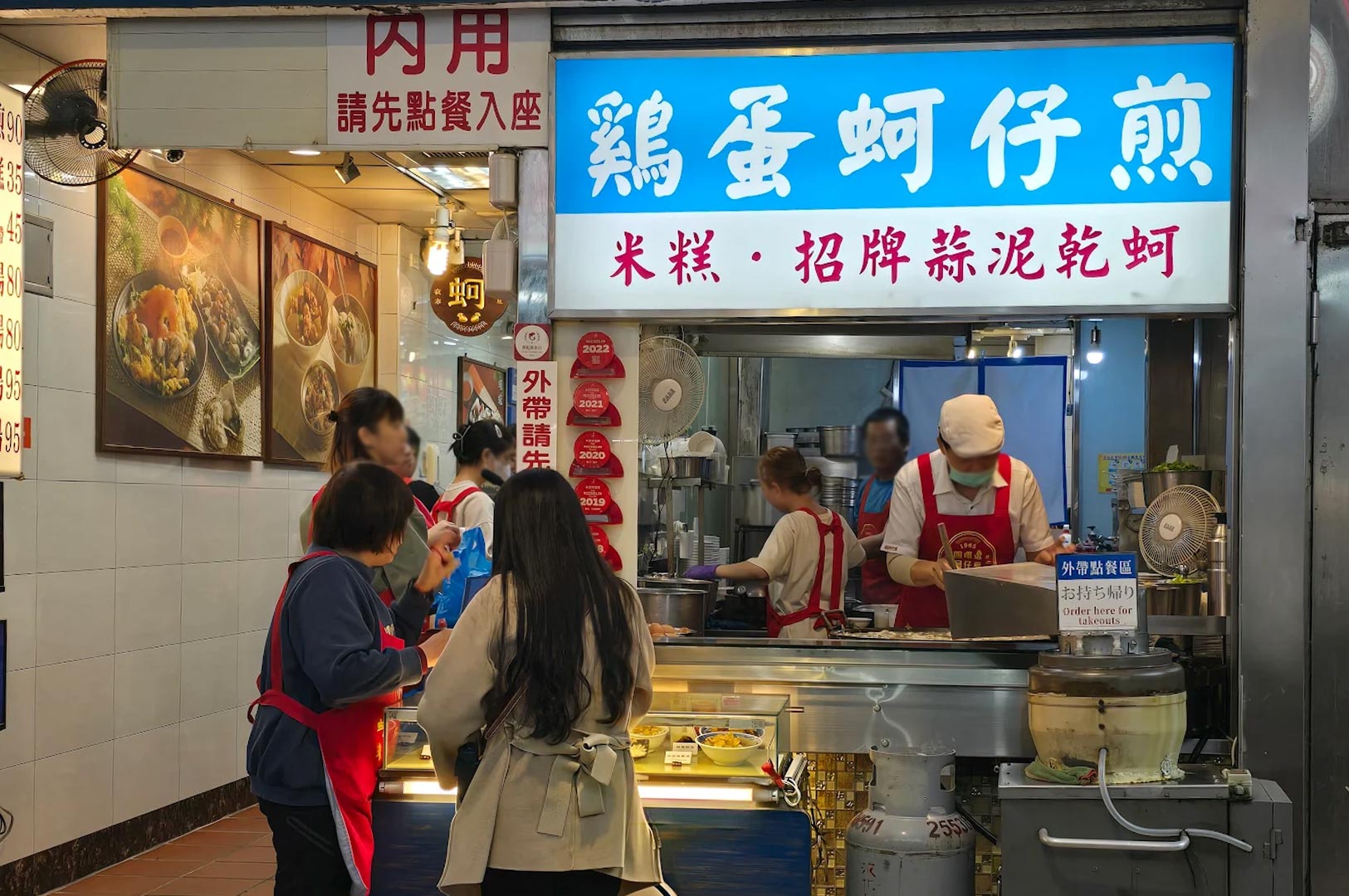 The storefront of the Michelin-recognized Yuan Huan Pien Oyster Omelet stall, showing staff in red aprons preparing food for a queue of customers.