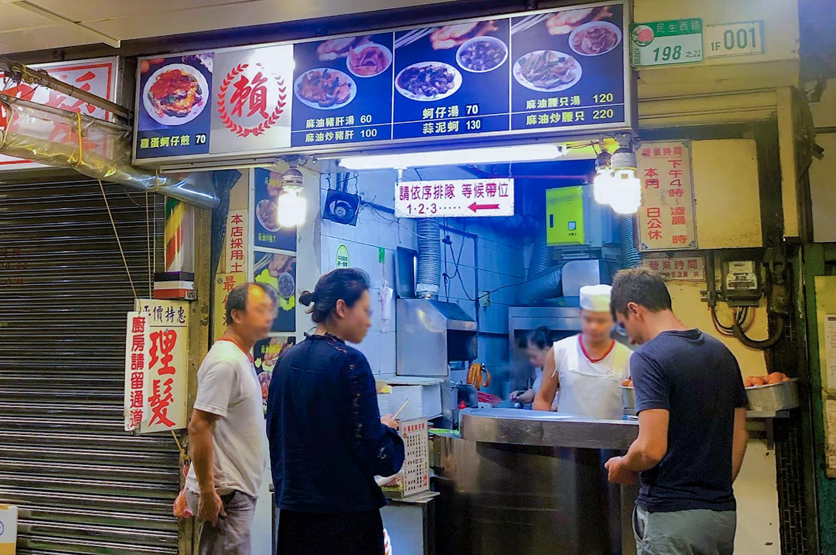 A traditional Taiwanese street food stall with a blue menu board displaying prices for oyster omelets and liver soups, with customers waiting at the counter.