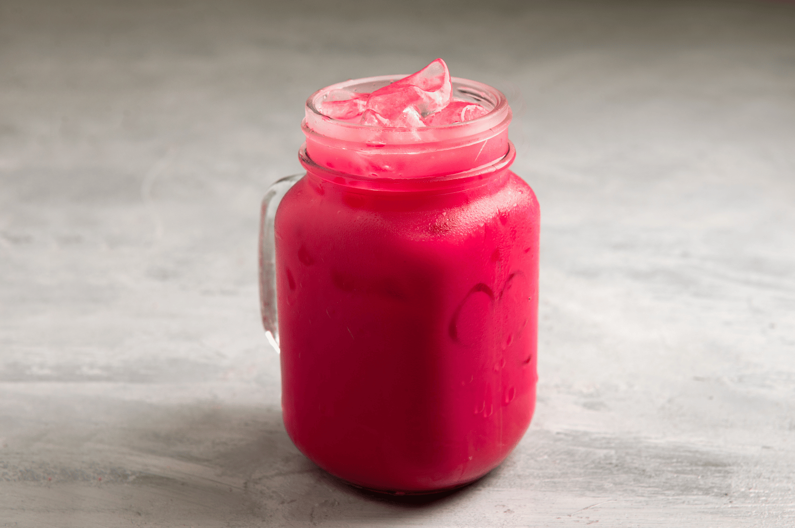 Vibrant pink iced Bandung in a mason jar with visible ice cubes, set against a neutral textured background—highlighting Singapore’s iconic rose syrup milk drink in its simplest, refreshing form.