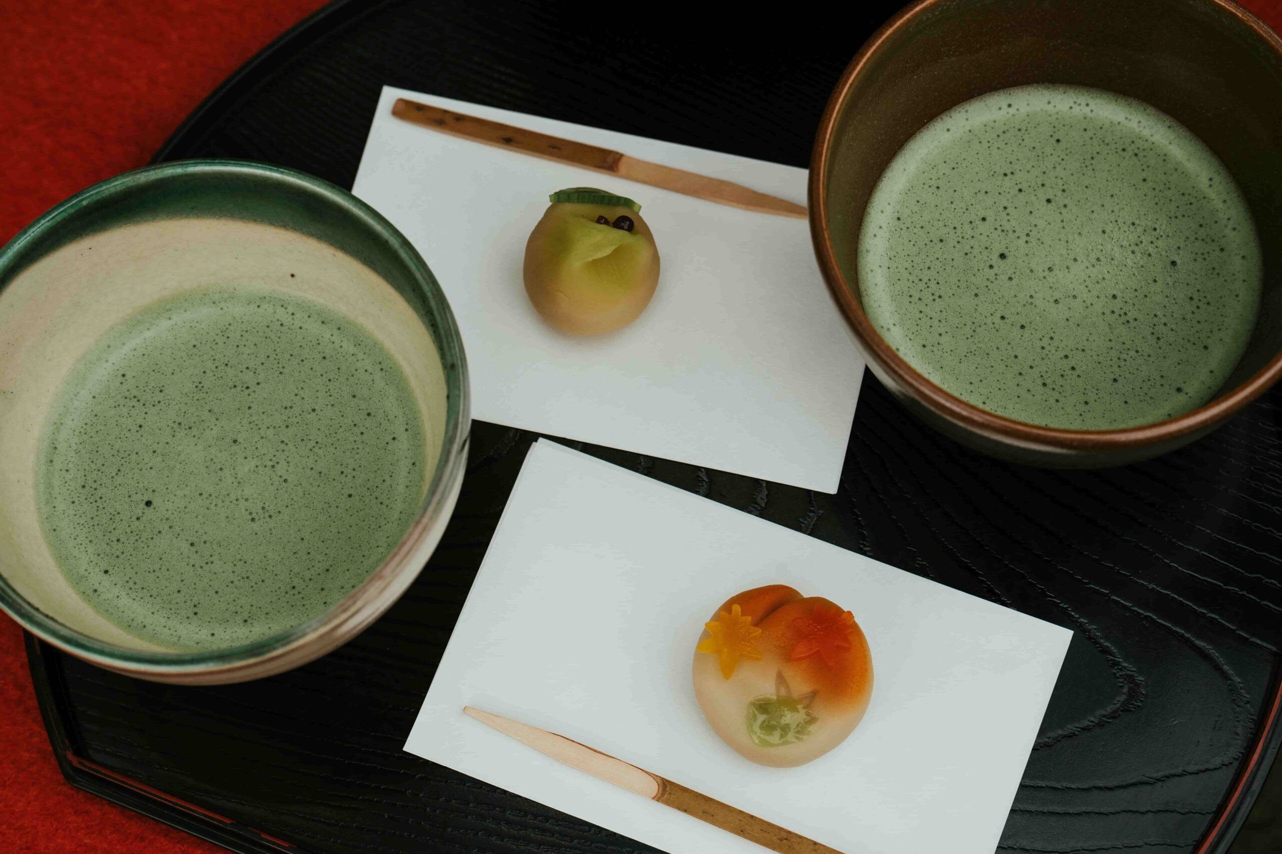Two pieces of seasonal wagashi sweets accompanied by two cups of matcha on a black tray.
