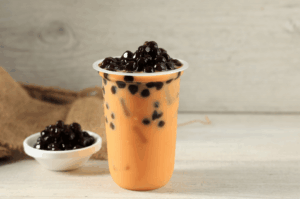 A plastic cup of bubble milk tea with black tapioca pearls, served alongside a small bowl of boba on a rustic wooden surface—an example of bubble tea’s colorful variations that spread worldwide from its Taiwanese origins.