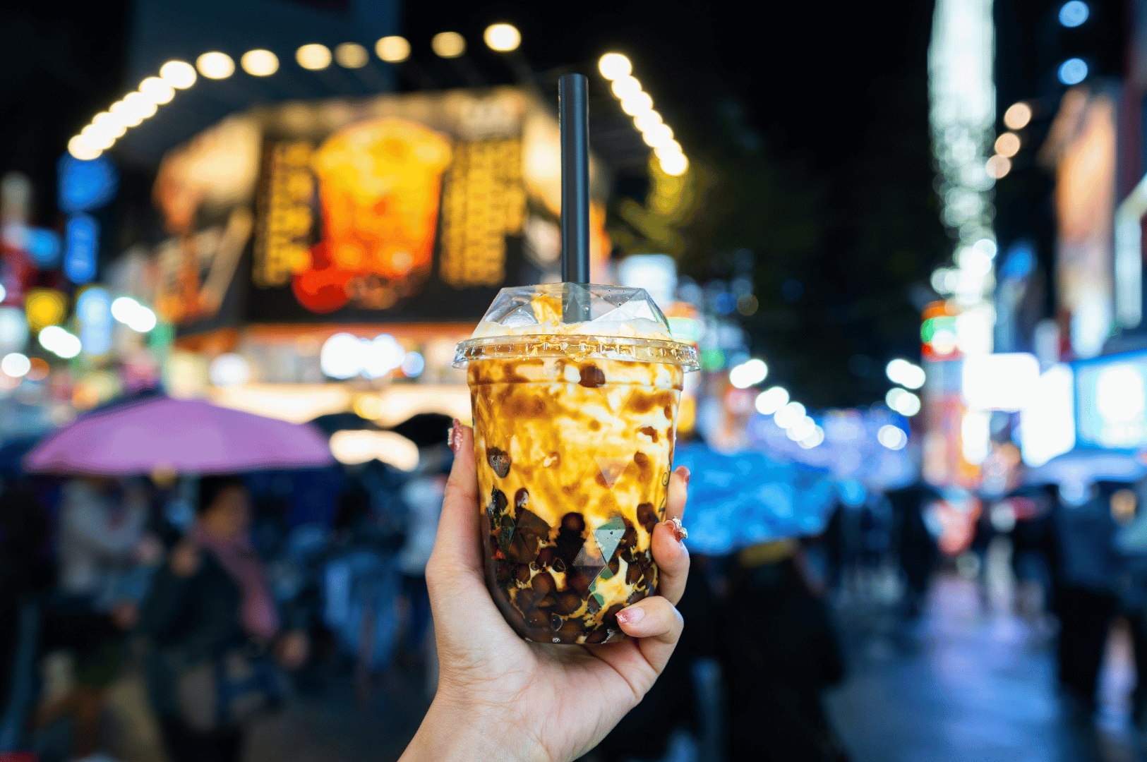 Close-up of brown sugar milk tea with boba, held against a neon-lit street filled with people and umbrellas, reflecting bubble tea’s rise from Taiwan to an international nightlife staple.