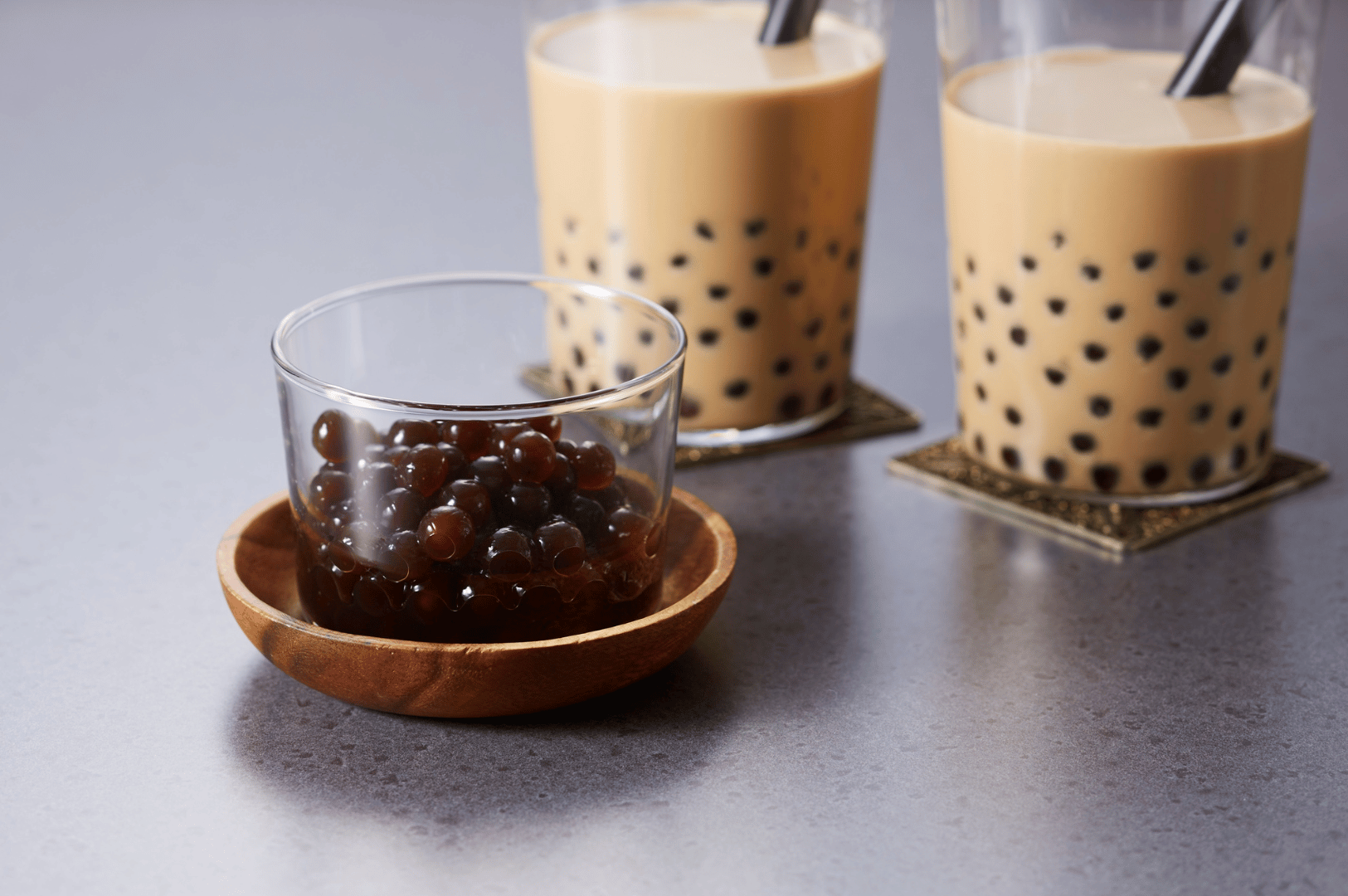 Two glasses of classic milk tea with tapioca pearls and spoons, served on ornate coasters beside a small bowl of boba, showcasing the essential ingredients of Taiwan’s bubble tea.