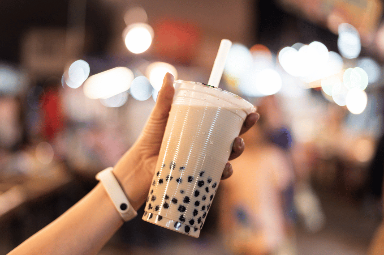 Hand holding a cup of milk tea with tapioca pearls in a lively street market setting, showing bubble tea’s roots in Taiwan’s street food culture and its worldwide popularity.