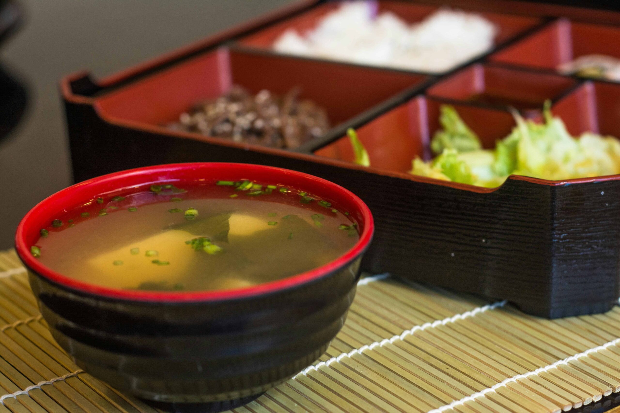 Food In Japan - 8 A bowl of Miso soup beside a bento box, placed over a bamboo mat.