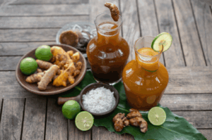 Rustic tabletop scene showcasing two glass carafes of homemade jamu, a traditional Indonesian herbal tonic. One carafe is garnished with lime, the other with turmeric root, surrounded by fresh ingredients—ginger, turmeric, limes, white rice, and a wooden mortar and pestle—arranged on a large green leaf. The setup evokes natural wellness, ancestral healing, and the modern revival of jamu as a holistic remedy.