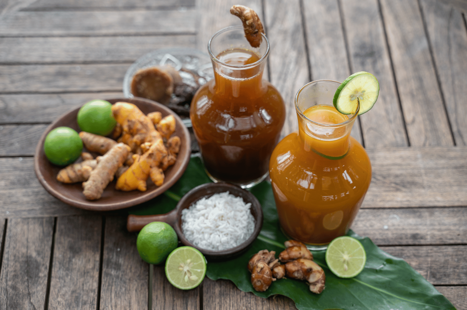 Rustic tabletop scene showcasing two glass carafes of homemade jamu, a traditional Indonesian herbal tonic. One carafe is garnished with lime, the other with turmeric root, surrounded by fresh ingredients—ginger, turmeric, limes, white rice, and a wooden mortar and pestle—arranged on a large green leaf. The setup evokes natural wellness, ancestral healing, and the modern revival of jamu as a holistic remedy.