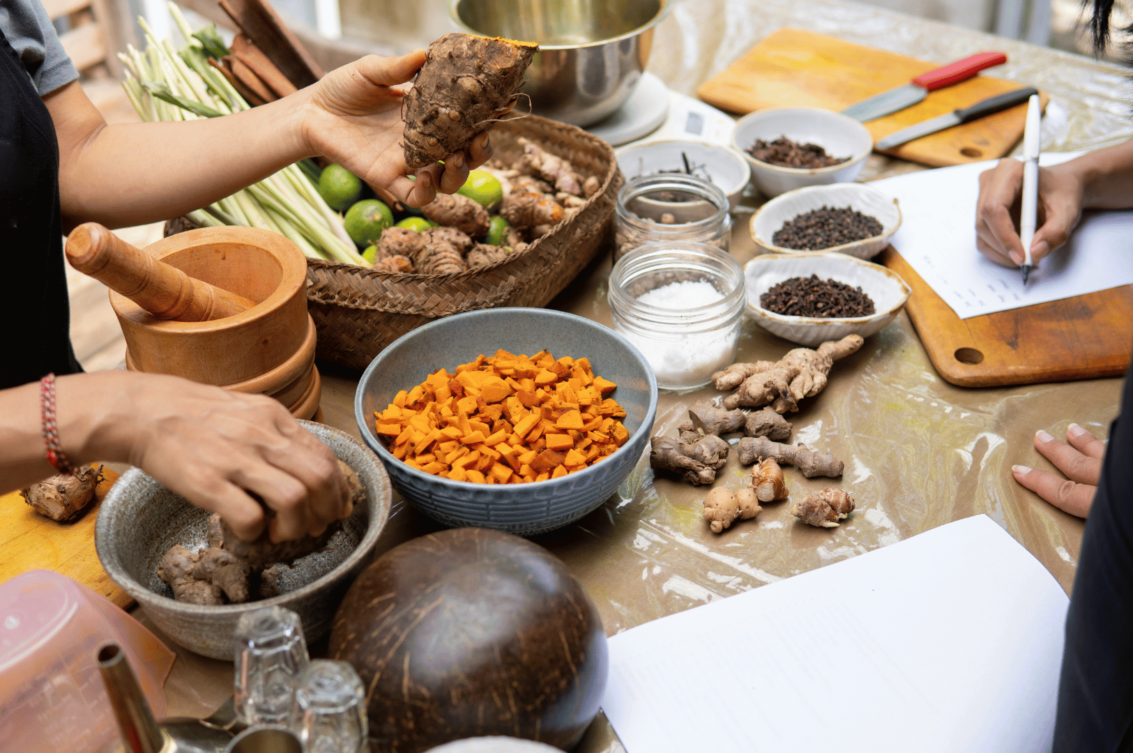 Hands-on jamu preparation scene featuring fresh turmeric, ginger, lemongrass, limes, and traditional spices like cloves and peppercorns arranged on a rustic table. Multiple hands chop, grind, and take notes, evoking a communal, educational setting rooted in ancestral Indonesian wellness practices. Mortar and pestle, coconut bowls, and handwritten recipes highlight the fusion of tradition and modern rediscovery.