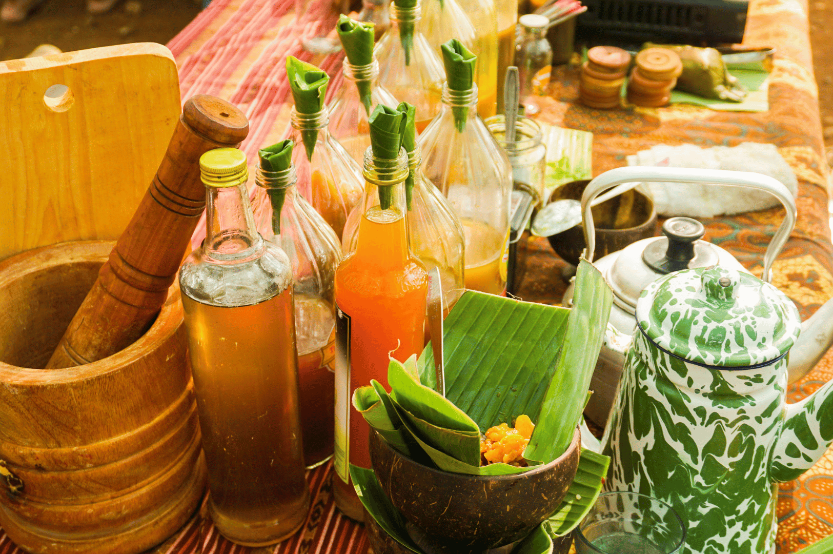 Traditional jamu preparation setup featuring glass bottles of herbal infusions capped with banana leaves, surrounded by coconut bowls, a wooden mortar and pestle, and enamel kettles. Yellow turmeric paste and natural ingredients rest on banana leaves, evoking ancestral Indonesian wellness rituals and the sensory richness of plant-based healing.