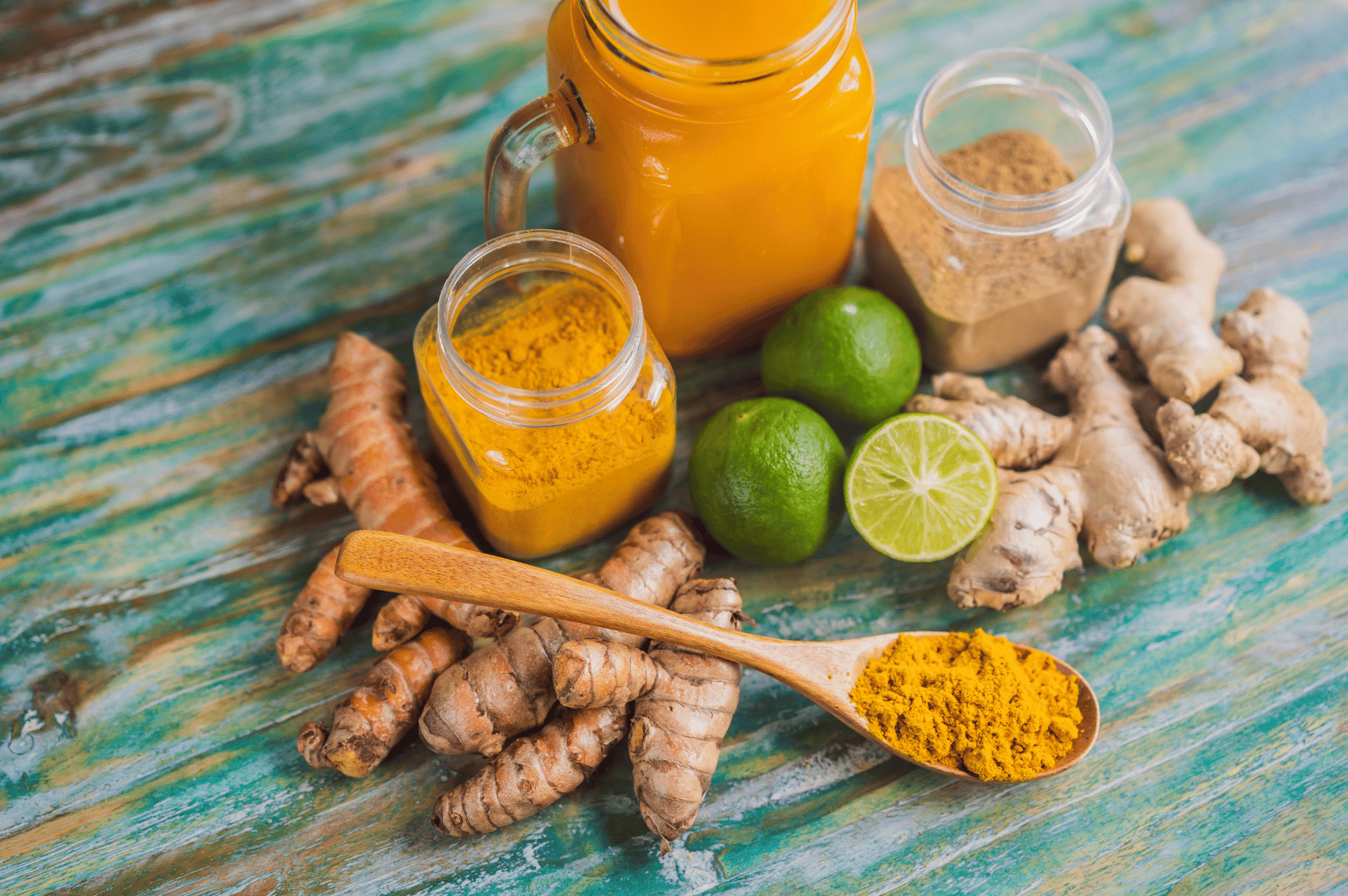 Flat lay of jamu ingredients on a rustic wooden surface: fresh turmeric and ginger roots, whole and halved limes, ground turmeric in a wooden spoon and glass jar, a second jar of brown spice, and a mason jar of vibrant turmeric juice. The arrangement highlights the raw, healing elements of traditional Indonesian herbal medicine.