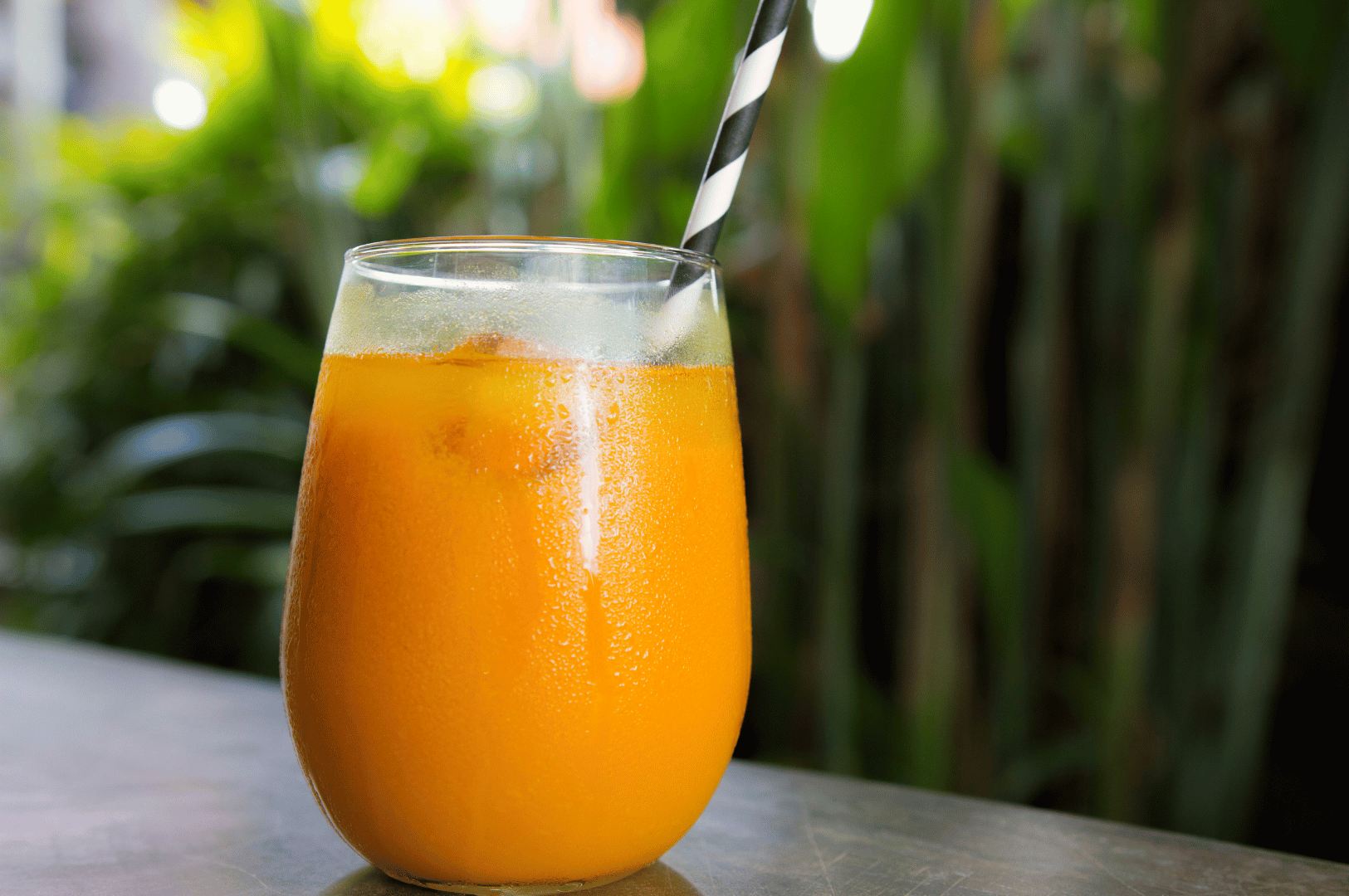 Chilled glass of vibrant orange jamu with ice cubes and a striped straw, set against a lush tropical backdrop. Condensation glistens on the glass, evoking refreshment and relaxation. The garden setting and vivid color celebrate jamu’s evolution from ancestral remedy to modern wellness ritual.