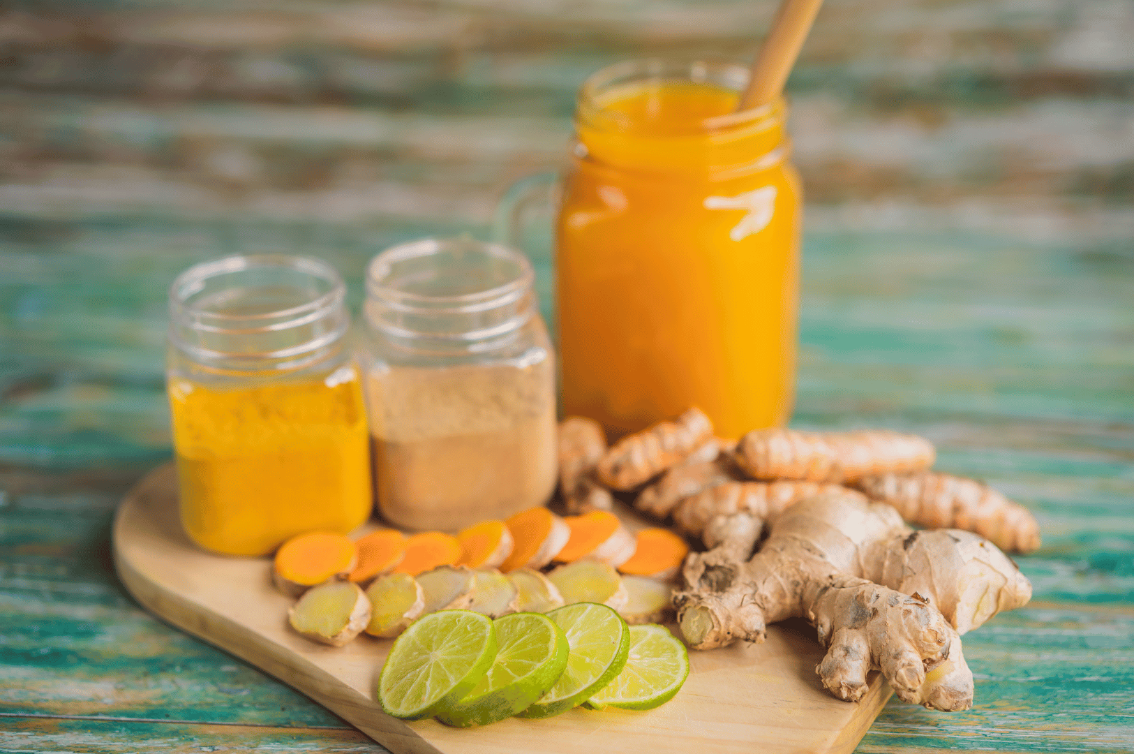 Wooden cutting board with fresh slices of lime, turmeric, and ginger alongside their powdered forms in glass jars. A large jar of bright orange jamu with a wooden stirrer anchors the scene, set against a teal-hued rustic backdrop. The composition highlights the transformation of raw ingredients into a vibrant wellness drink rooted in Indonesian tradition.