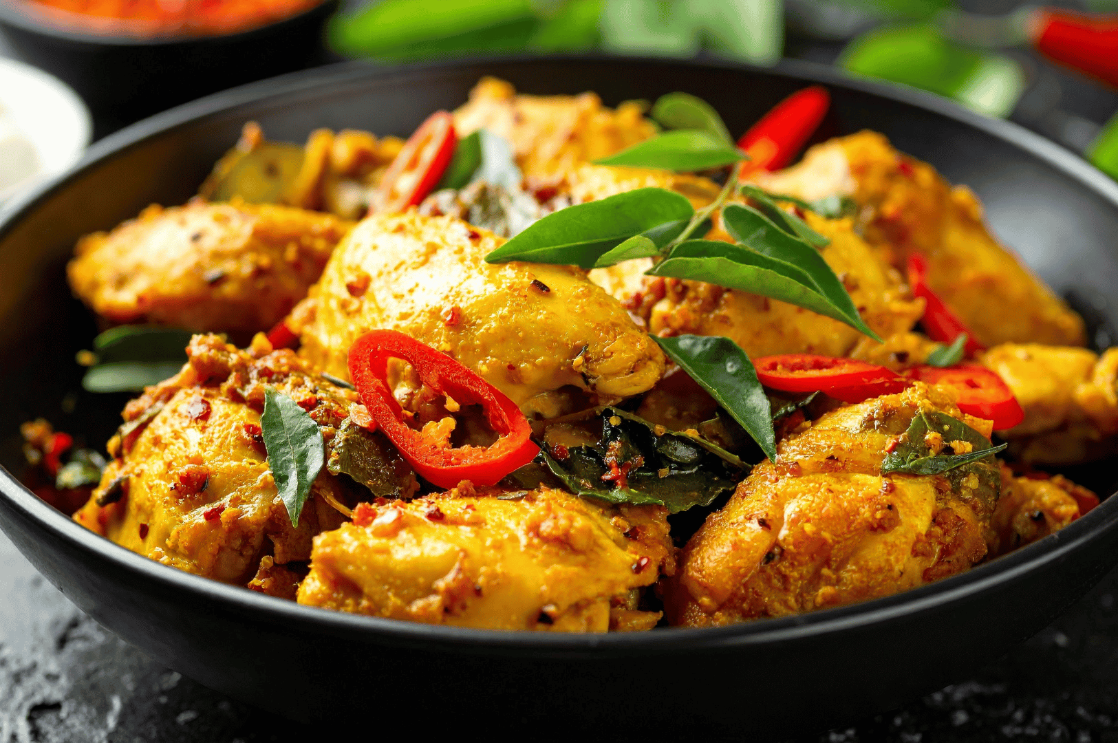 Close-up of a slow-cooked South Asian chicken curry in a black bowl, topped with red chili slices and fresh curry leaves on a dark background.