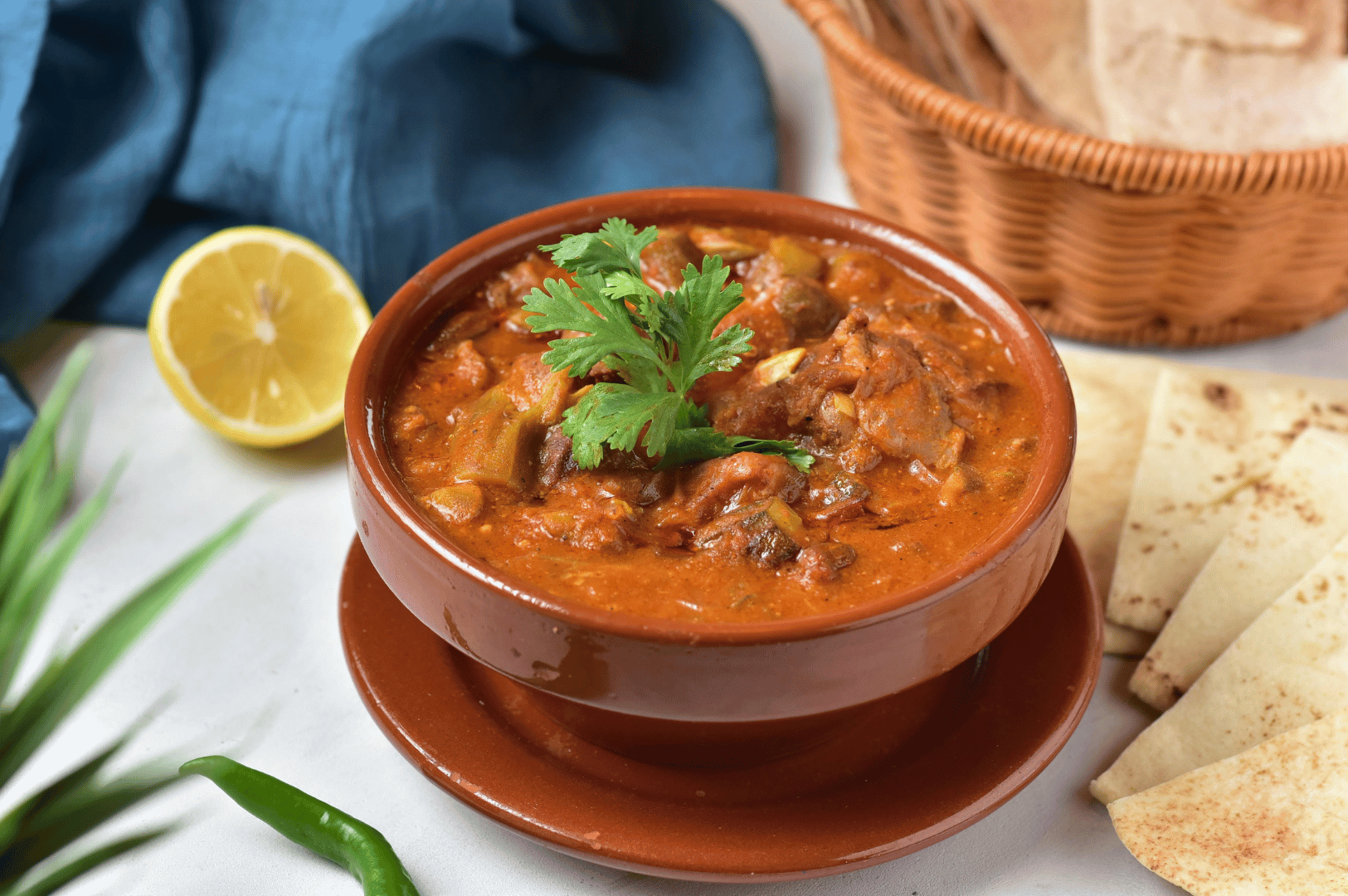 Close-up of hearty chicken curry in a clay bowl, thick tomato gravy and herbs, with naan and lemon on the side—showing the warmth of time-honored cooking.