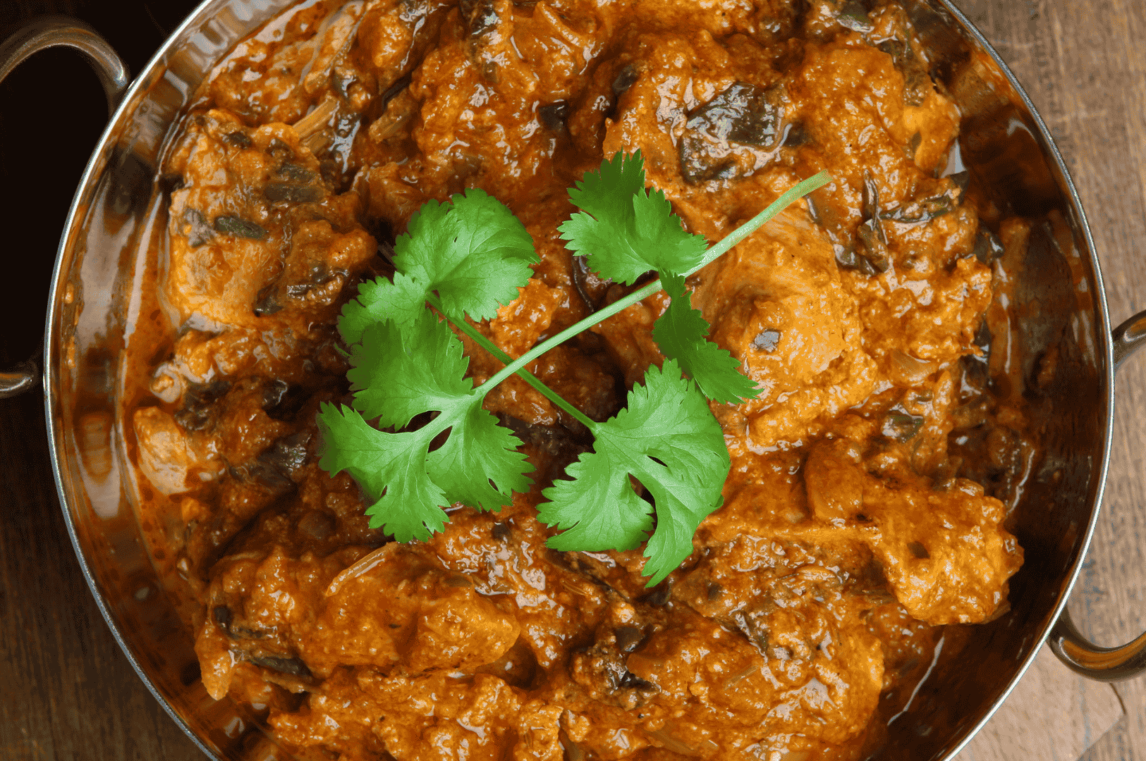 Overhead view of a rich, slow-simmered chicken curry in a metal bowl, garnished with fresh cilantro—showcasing the deep color and texture of long-cooked spices