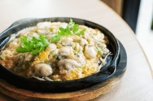 A sizzling Taiwanese oyster omelet served in a black cast iron skillet atop a wooden base, featuring plump oysters nestled in a golden, crispy-edged egg and starch batter. Garnished with fresh cilantro and captured mid-steam, the dish evokes street food nostalgia and culinary warmth, set against a rustic restaurant backdrop.