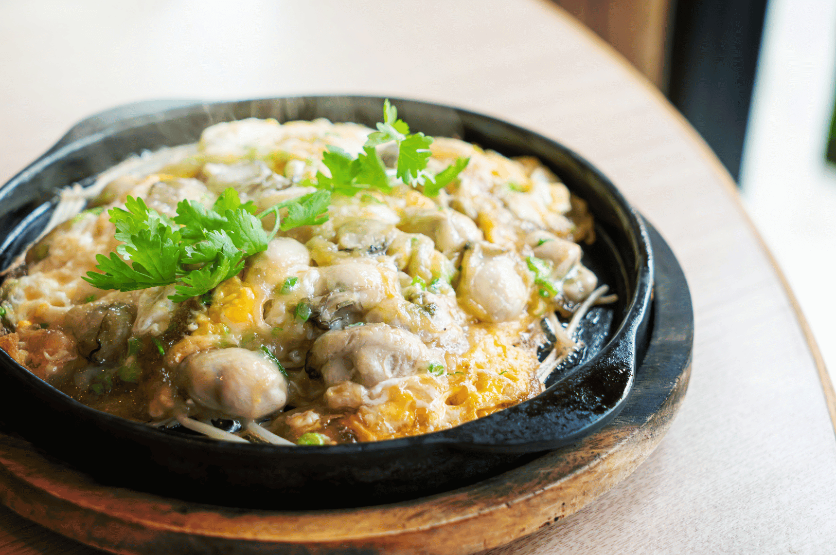 A sizzling Taiwanese oyster omelet served in a black cast iron skillet atop a wooden base, featuring plump oysters nestled in a golden, crispy-edged egg and starch batter. Garnished with fresh cilantro and captured mid-steam, the dish evokes street food nostalgia and culinary warmth, set against a rustic restaurant backdrop.