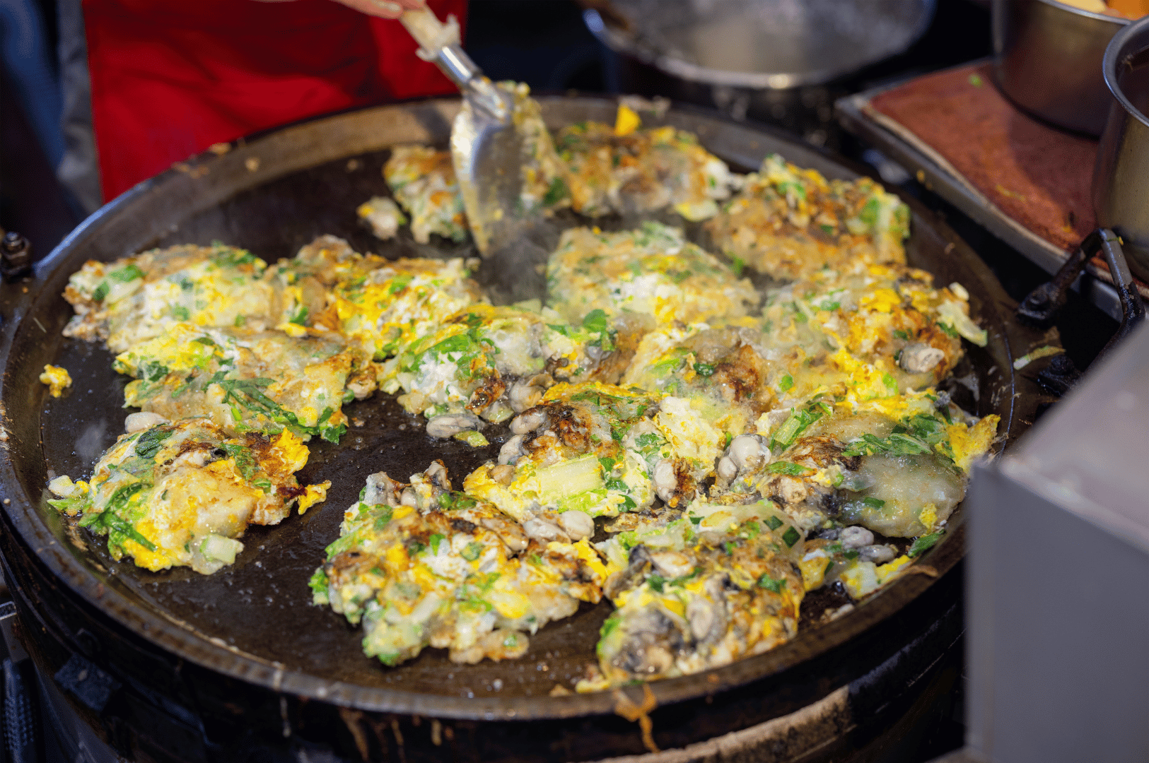 Multiple Taiwanese oyster omelets sizzling on a large round griddle at a bustling food stall, each golden brown and studded with oysters, chopped greens, and egg. A metal spatula lifts one mid-flip as steam rises from the hot surface, capturing the dynamic energy and street-side craftsmanship behind this beloved night market staple.