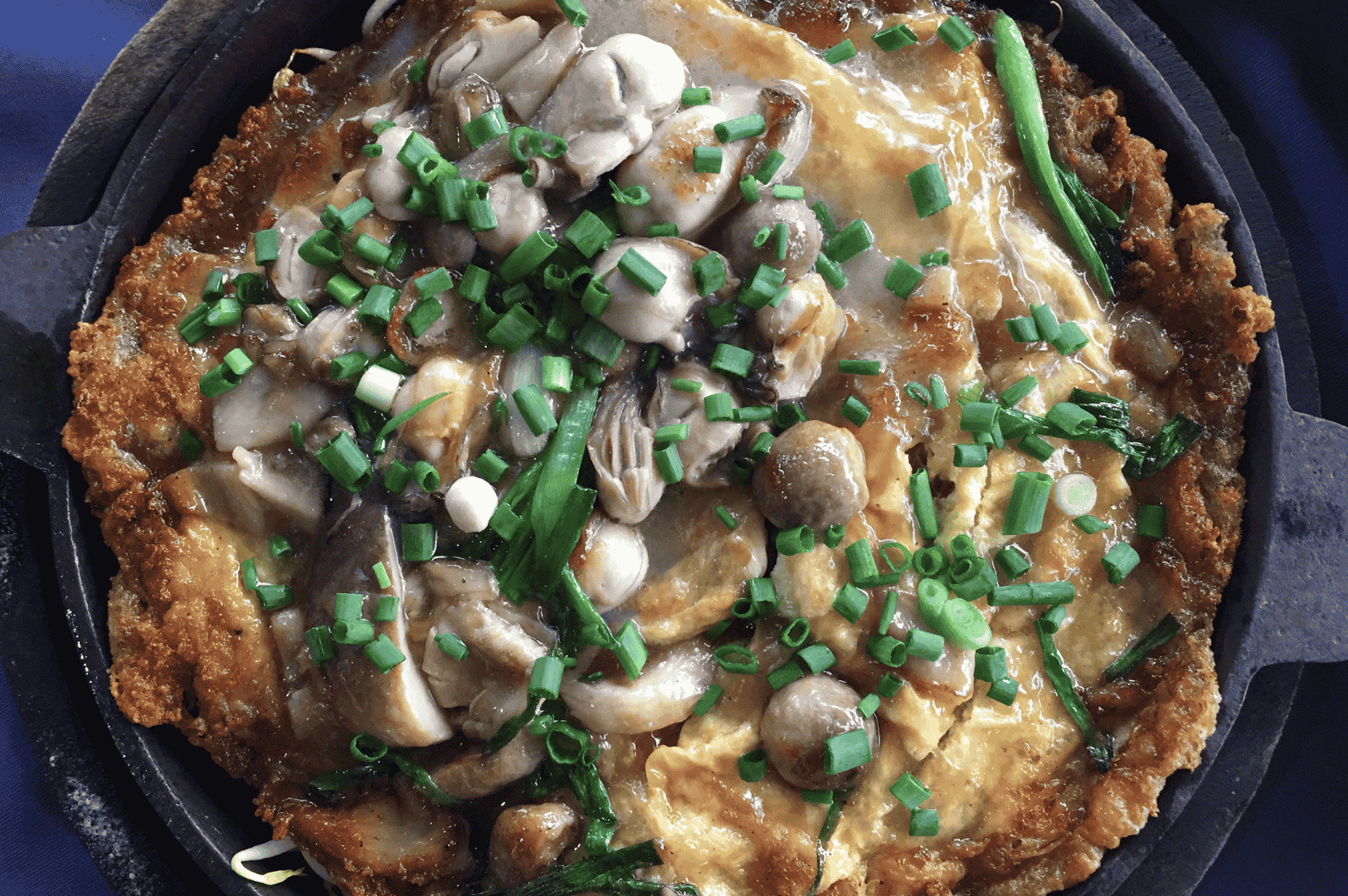 A Taiwanese oyster omelet served hot in a black cast iron skillet, layered with tender oysters, sautéed mushrooms, and chopped green onions over a crisp, golden egg base. The glossy sauce glistens under warm lighting, while the skillet presentation evokes rustic comfort and elevates the dish’s street food origins.