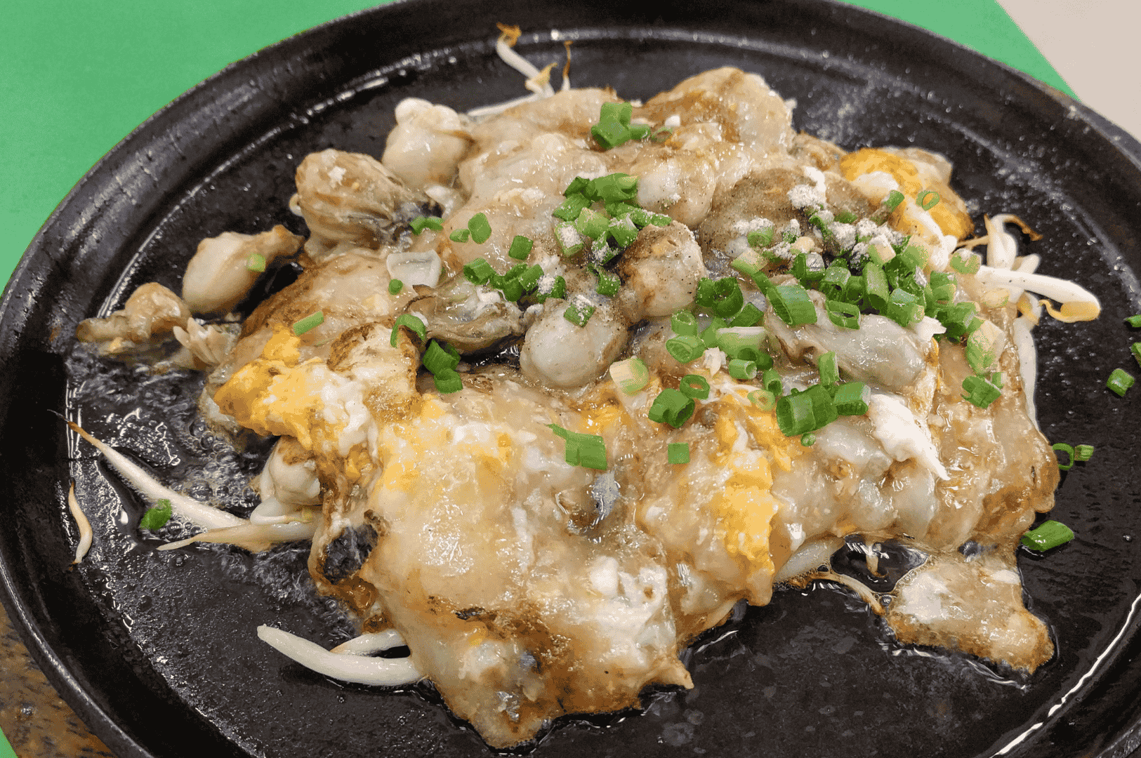 A freshly cooked Taiwanese oyster omelet sizzling on a black hot plate, featuring plump oysters, eggs, and a glossy, gelatinous batter layered over crisp bean sprouts. Garnished with chopped green onions and enveloped in rising steam, the dish captures the tactile contrast and savory depth that define this beloved night market classic.