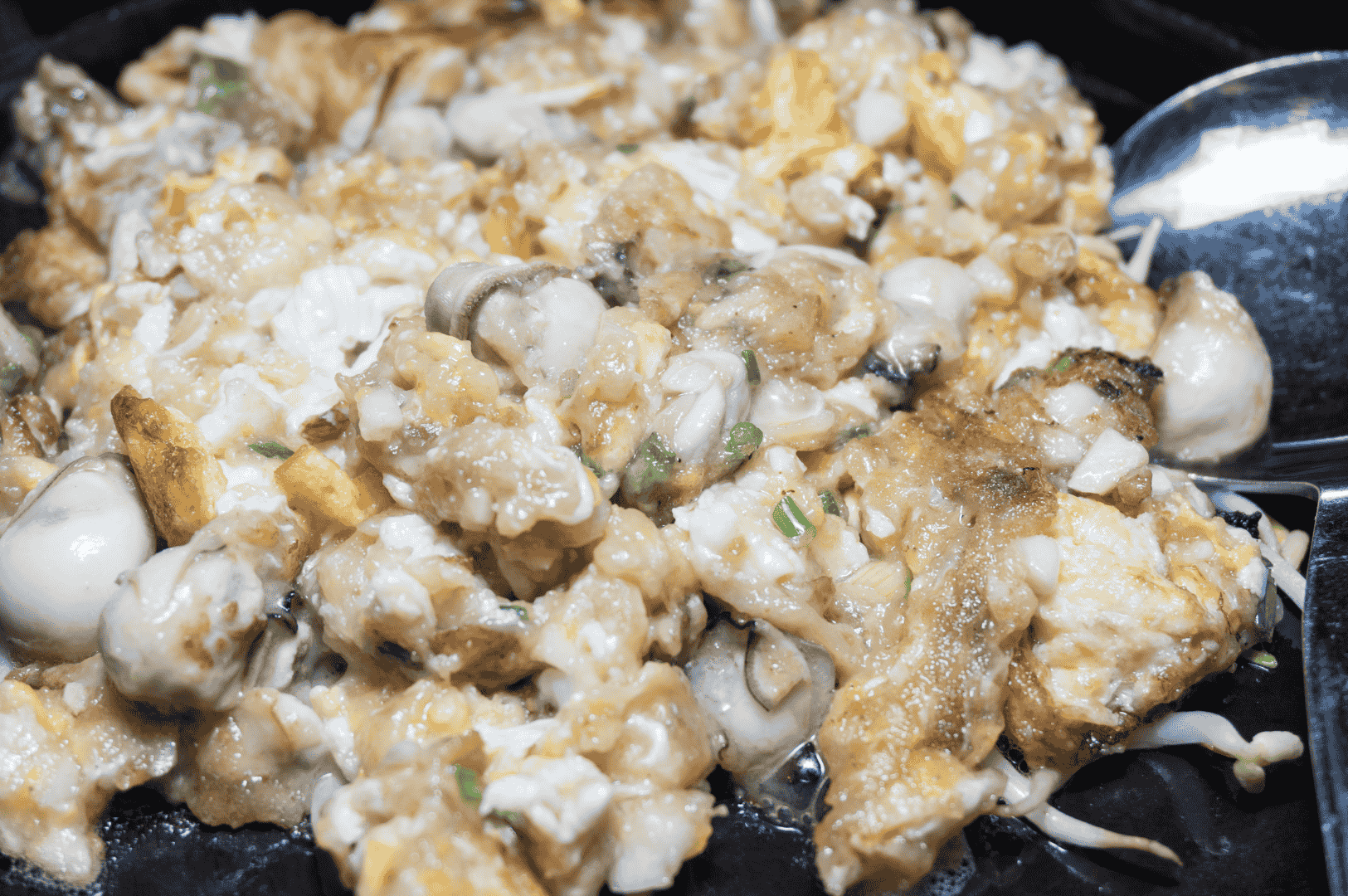 Close-up of a Taiwanese oyster omelet showcasing its thick, gelatinous texture with visible oysters, scrambled eggs, and scallion garnish. The edges are slightly crisp, and a metal spoon rests beside the dish, suggesting it’s ready to be enjoyed. The intimate framing highlights the dish’s layered textures and savory appeal, inviting the viewer into the moment of indulgence.