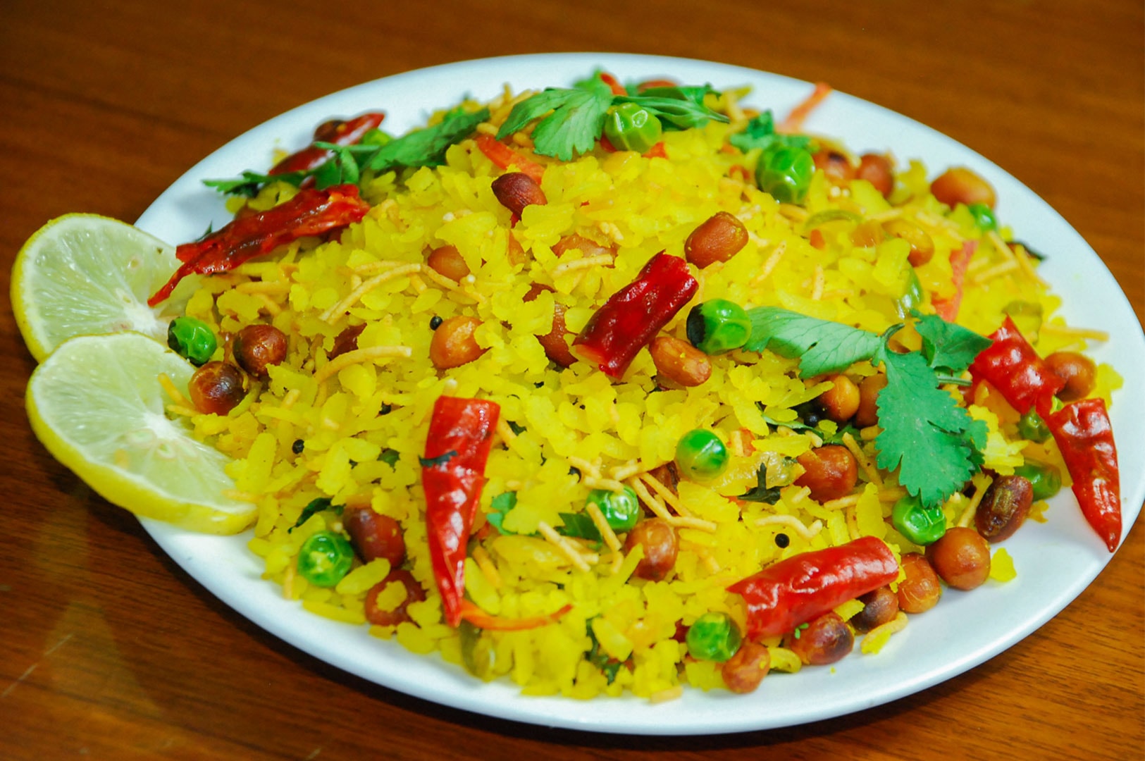 A plate of yellow Poha (flattened rice) cooked with peanuts, green peas, and red chilies, garnished with fresh cilantro and lemon slices.