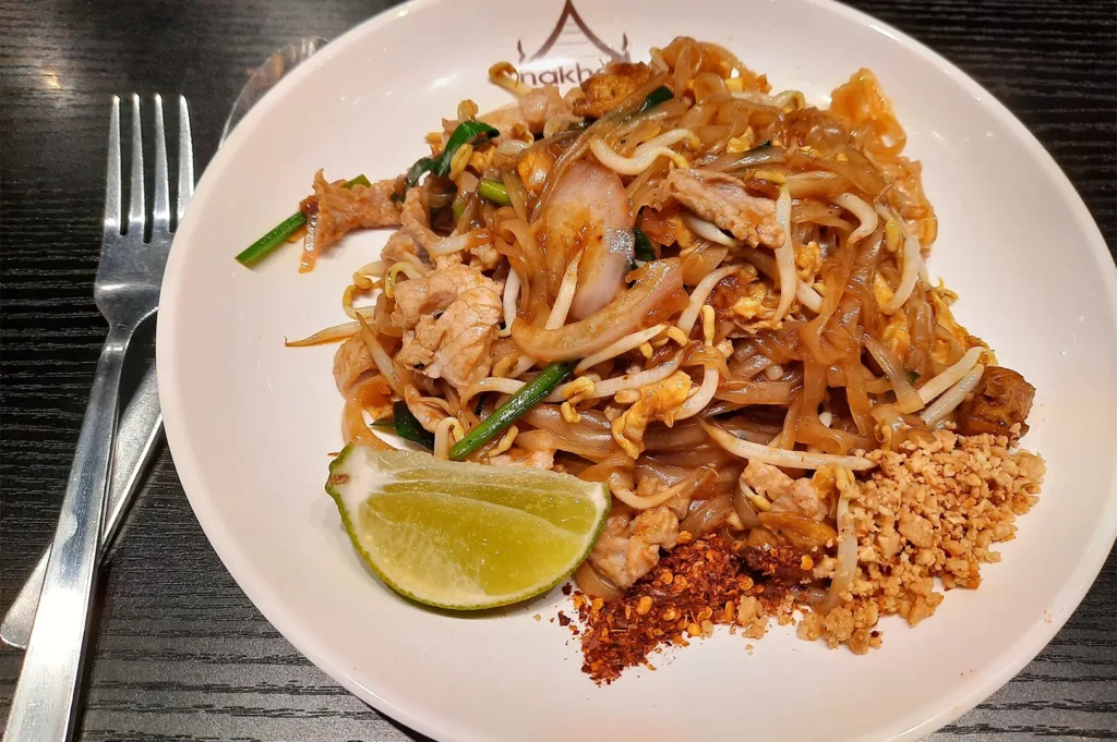Authentic Pad Thai with pork, rice noodles, bean sprouts, crushed peanuts, and chili flakes served with a lime wedge on a white plate.