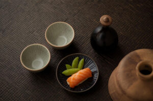 Flatlay composition of two small ceramic sake cups, a sake bottle, and a piece of salmon nigiri sushi with two snow peas on a small black plate.