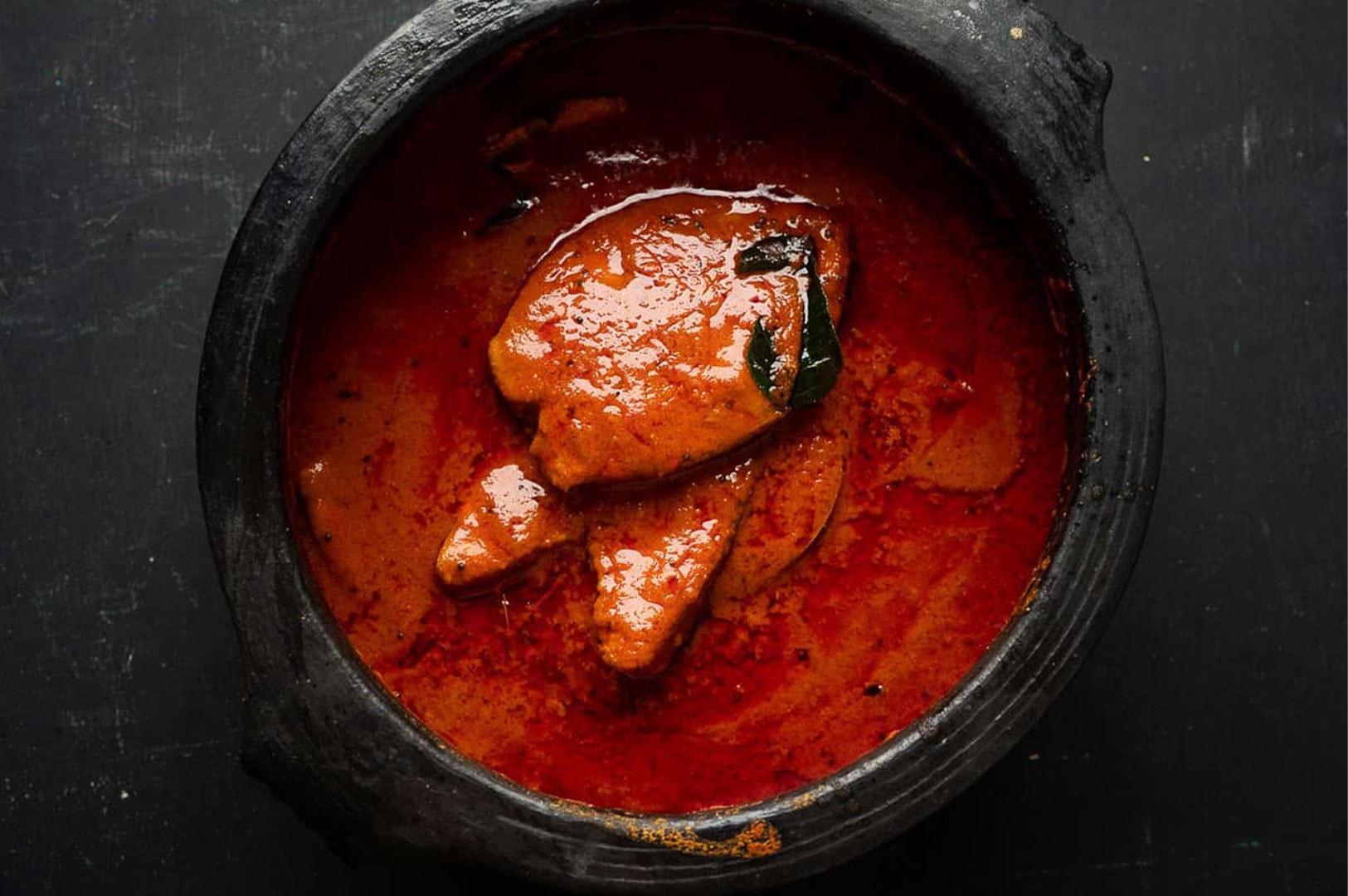 Traditional red fish curry served in a black earthenware clay pot (manchatti), highlighting the rich, spicy oil on the surface of the gravy.
