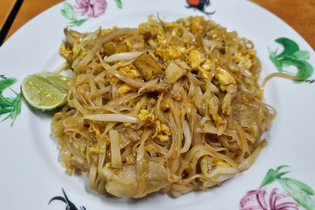Stir-fried Chicken Pad Thai with flat rice noodles, scrambled egg, tofu, and bean sprouts served on a floral patterned plate.