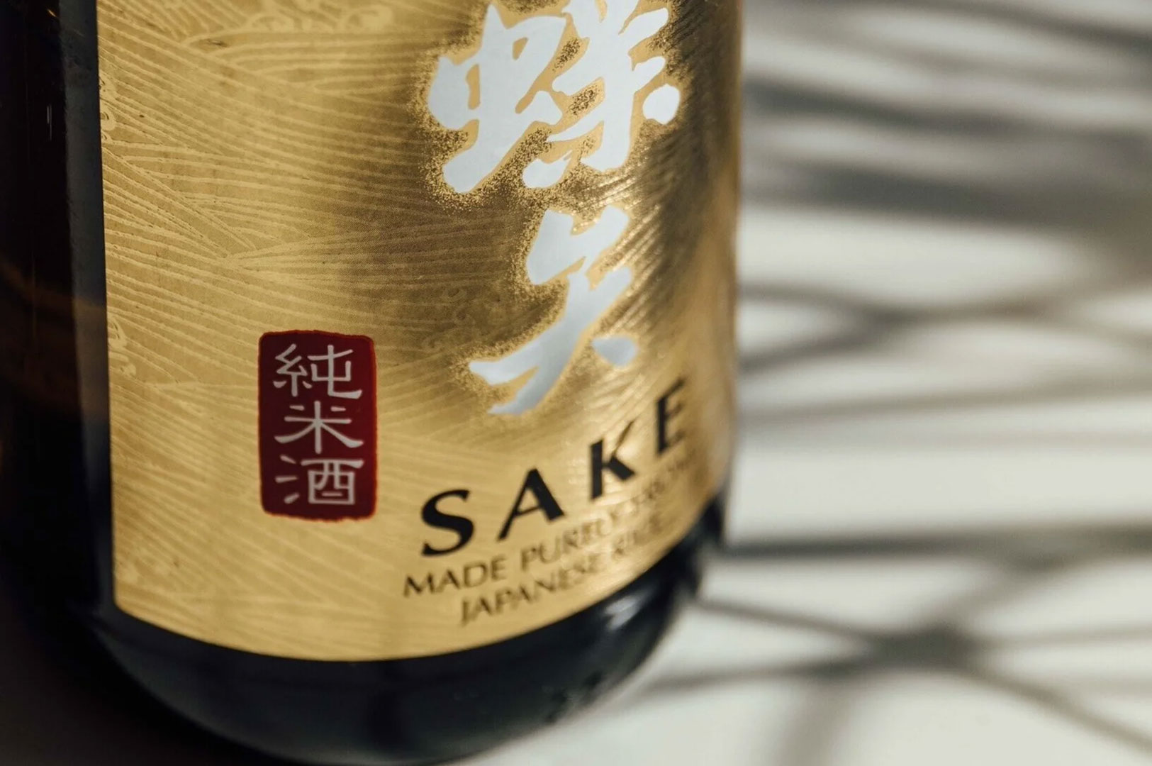 3-junmai-sake-bottle-gold-label-close-up Extreme close-up of a sake bottle label, featuring large white Japanese characters and the text "SAKE MADE PURELY FROM JAPANESE RICE" with the Junmai-shu stamp.