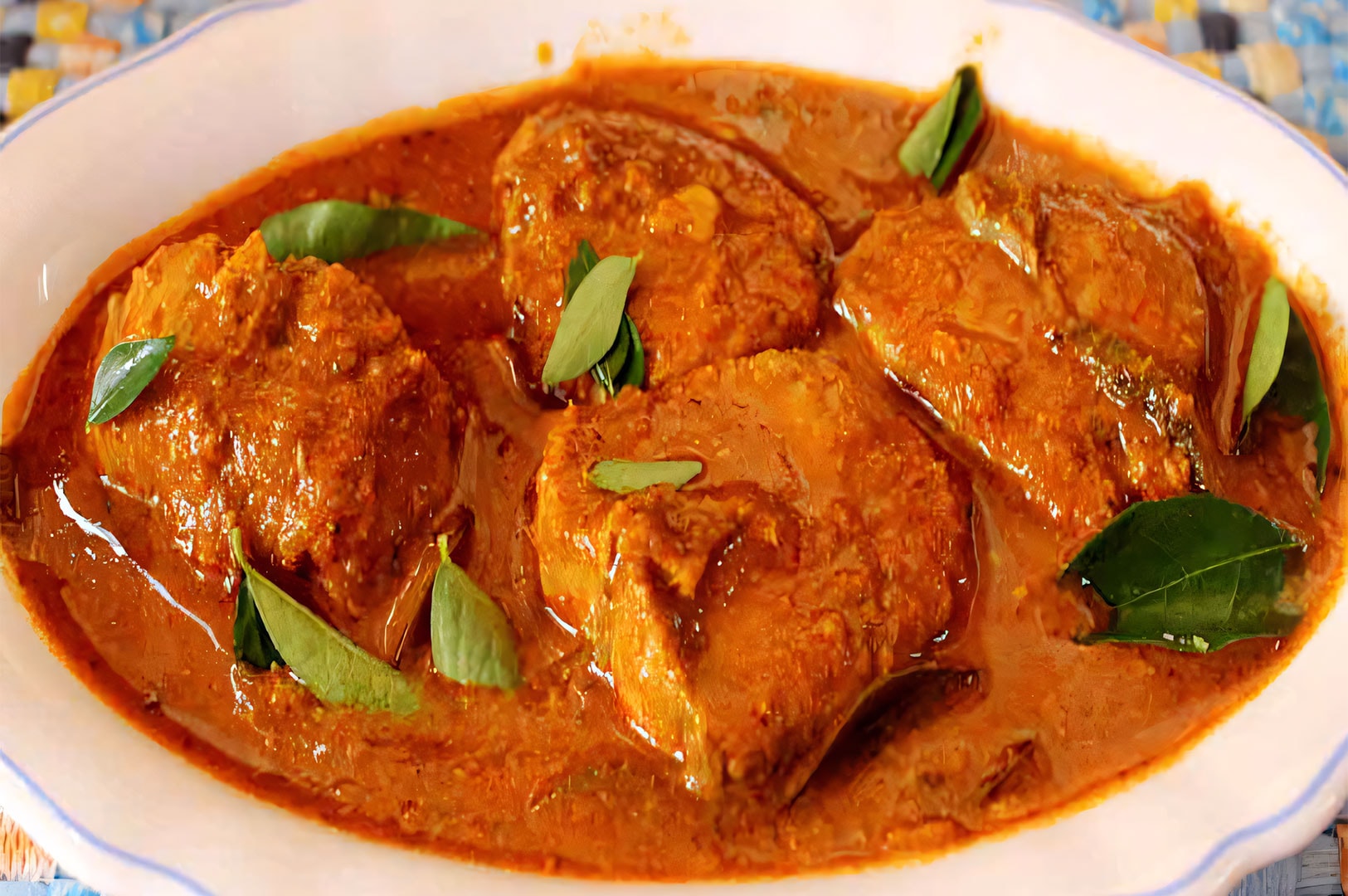 A white serving dish filled with spicy Indian fish curry in a rich red gravy, garnished with fresh green curry leaves.