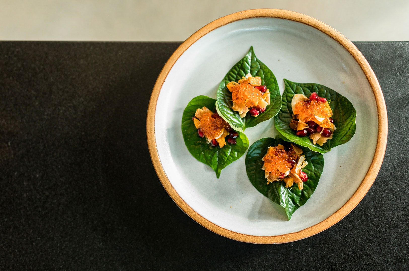 Four portions of Miang Kham, a traditional Thai appetizer, consisting of toasted coconut, ginger, peanuts, and other ingredients wrapped in fresh betel leaves and topped with orange roe, served on a ceramic plate.