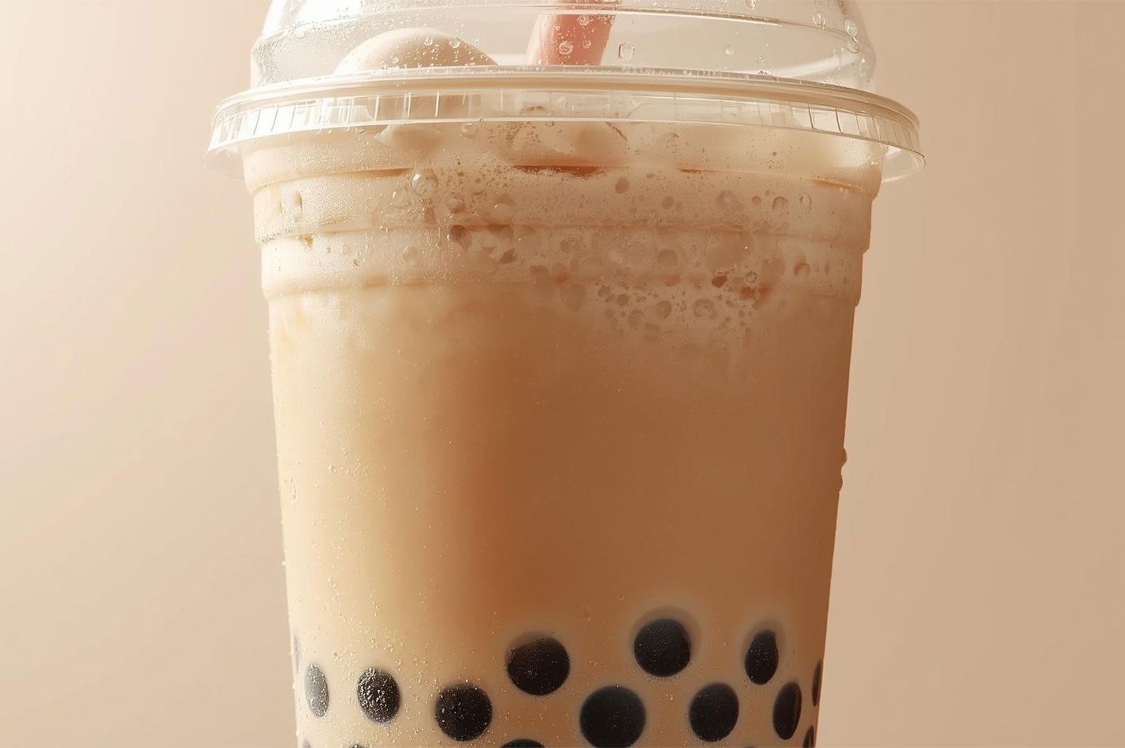 Close-up detail of a cold classic milk tea in a clear plastic takeaway cup, showing condensation droplets on the side and black tapioca pearls settled at the bottom.
