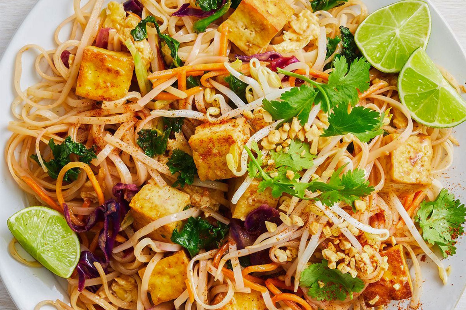 Overhead shot of a white plate filled with homemade Pad Thai noodles, featuring large cubes of fried tofu, fresh bean sprouts, and lime wedges.