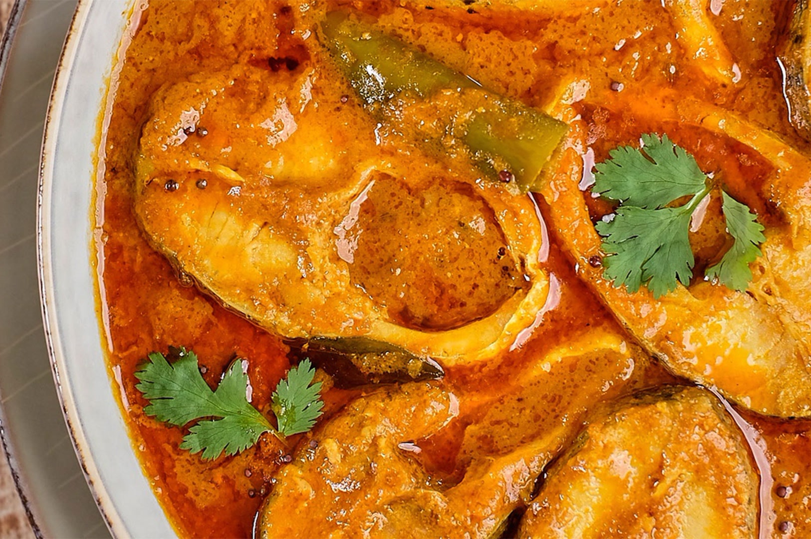 Close-up detail shot of a thick fish curry gravy showing the texture of the sauce, cooked green chilies, and fresh cilantro garnish.