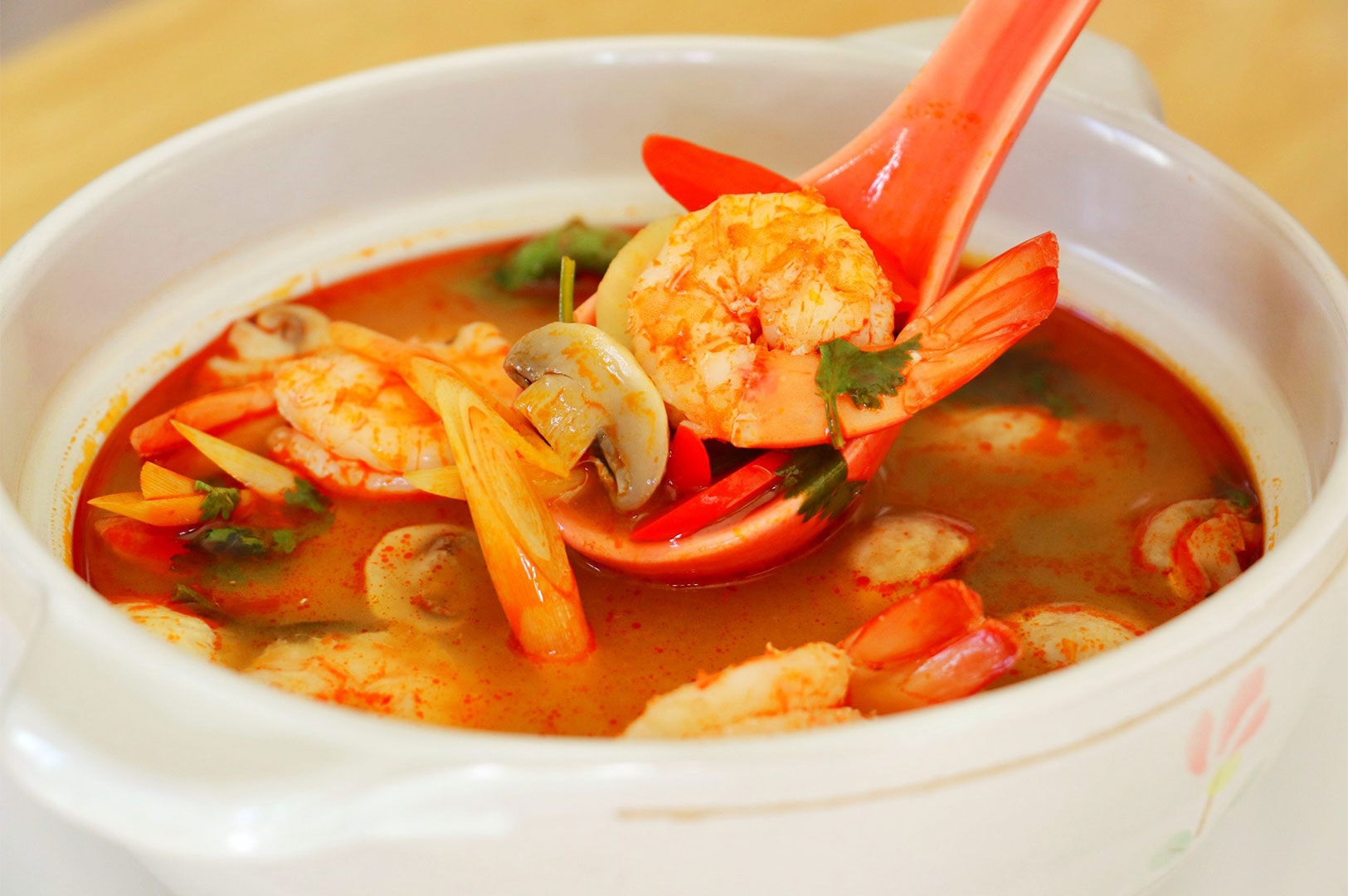 A close-up of a ladle scooping bright orange Tom Yum soup, showing a large shrimp, sliced mushrooms, and lemongrass, served in a white bowl.