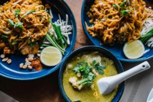 The image shows a close-up of a table setting with three dishes. Two blue plates contain what appears to be Pad Thai, garnished with bean sprouts, green onions, lime wedges, peanuts, and red pepper flakes.