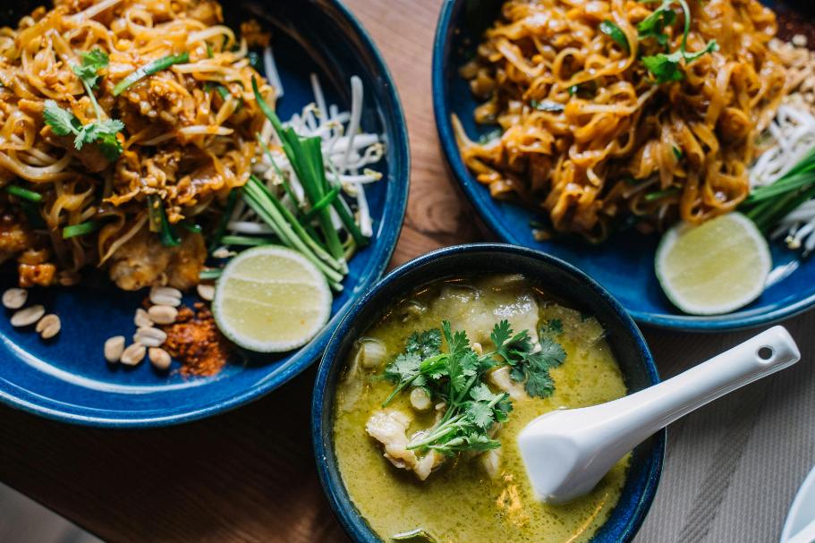 The image shows a close-up of a table setting with three dishes. Two blue plates contain what appears to be Pad Thai, garnished with bean sprouts, green onions, lime wedges, peanuts, and red pepper flakes.