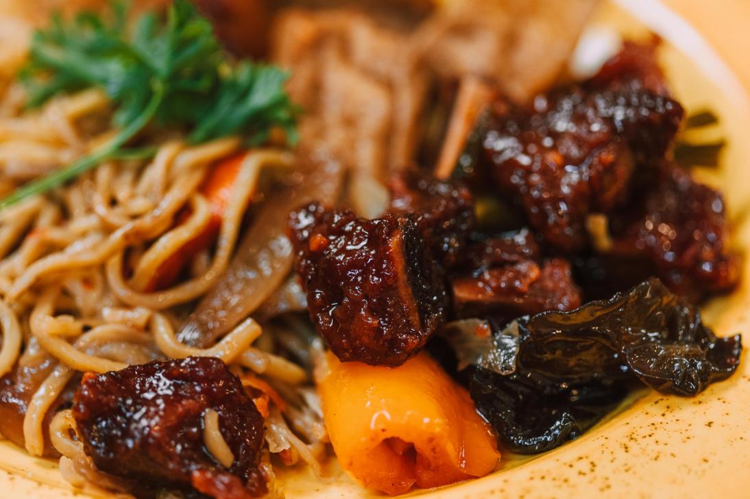 This close-up shot highlights a savory noodle dish featuring glossy, dark-glazed chunks of meat paired with vibrant orange bell peppers and dark vegetables.