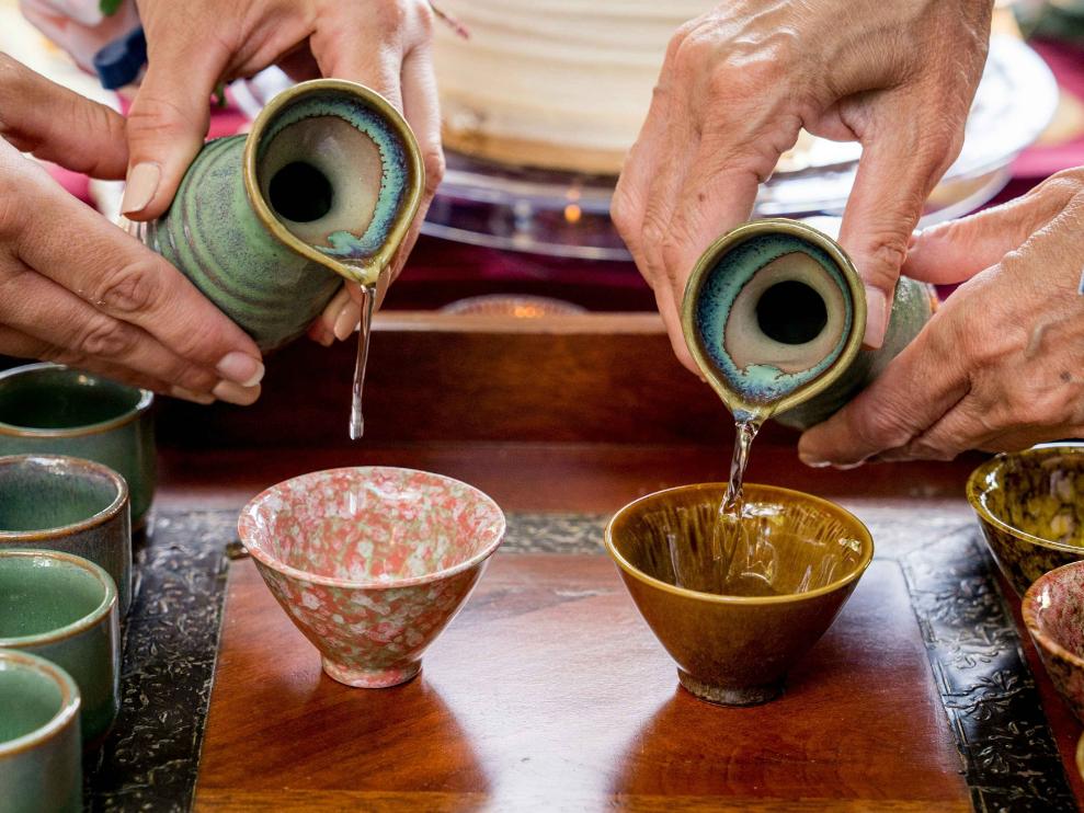 Understanding Sake - 2 An image of two Sake being poured into traditional Japanese ceramic cups on top of a vintage wooden table..