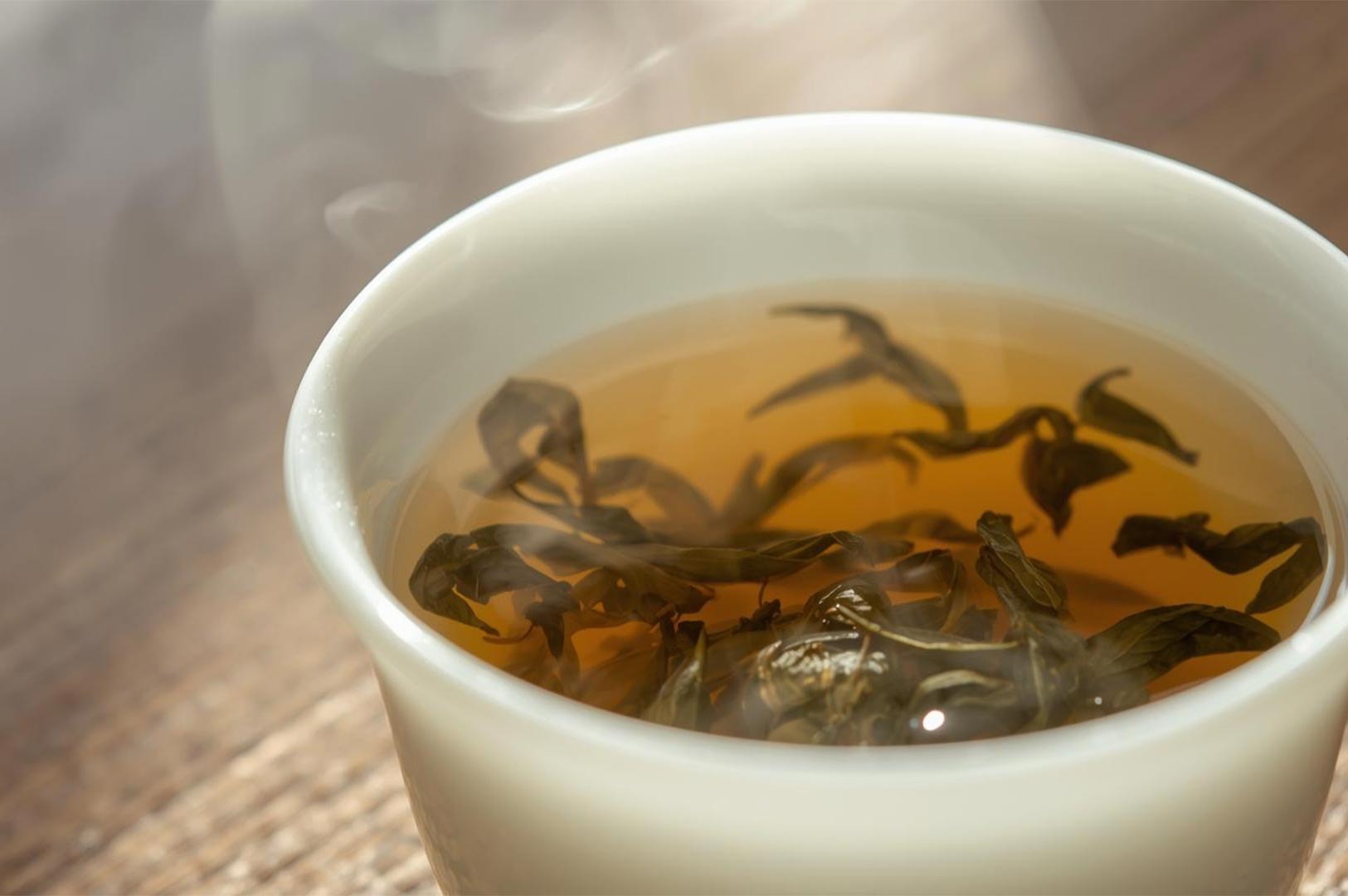 Close-up of hot, steaming oolong or herbal tea leaves steeping in an amber liquid inside a small white ceramic cup.