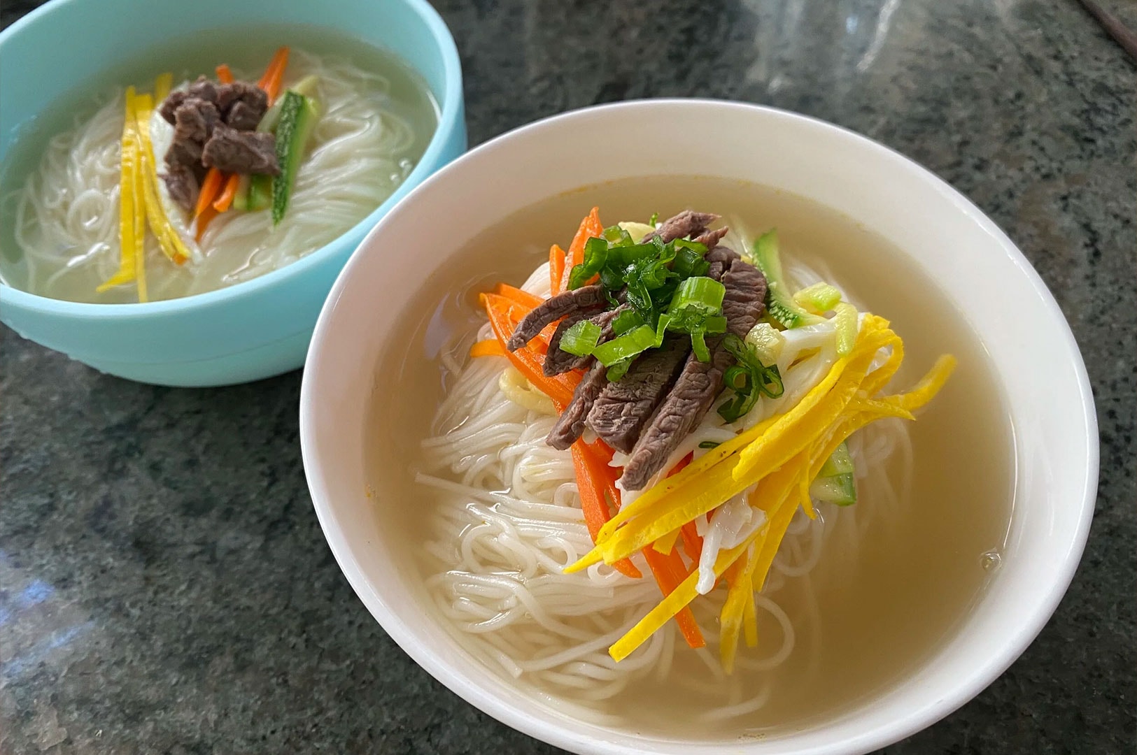 Two bowls of Janchi Guksu, a warm Korean noodle soup made with thin wheat noodles in a clear broth. The toppings include sliced beef, julienned yellow egg garnish, carrots, zucchini, and chopped green onions.