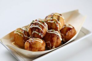 Six golden-brown takoyaki balls served in a traditional bamboo paper boat, drizzled with brown sauce and mayonnaise, and sprinkled with green dried seaweed (aonori).