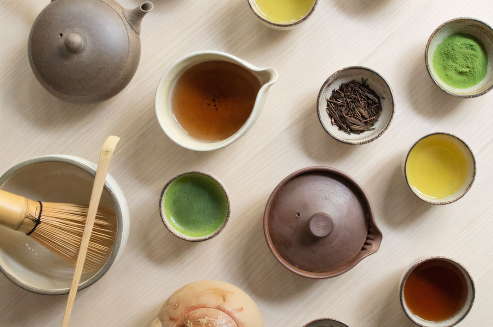 An overhead flat lay showing a variety of Japanese tea equipment, including a clay teapot, a bamboo matcha whisk, and various bowls containing matcha and roasted tea.