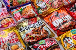 A colorful flat lay of various popular instant noodle brands, including Shin Ramyun, Buldak, Chapagetti, and Indomie, arranged together.